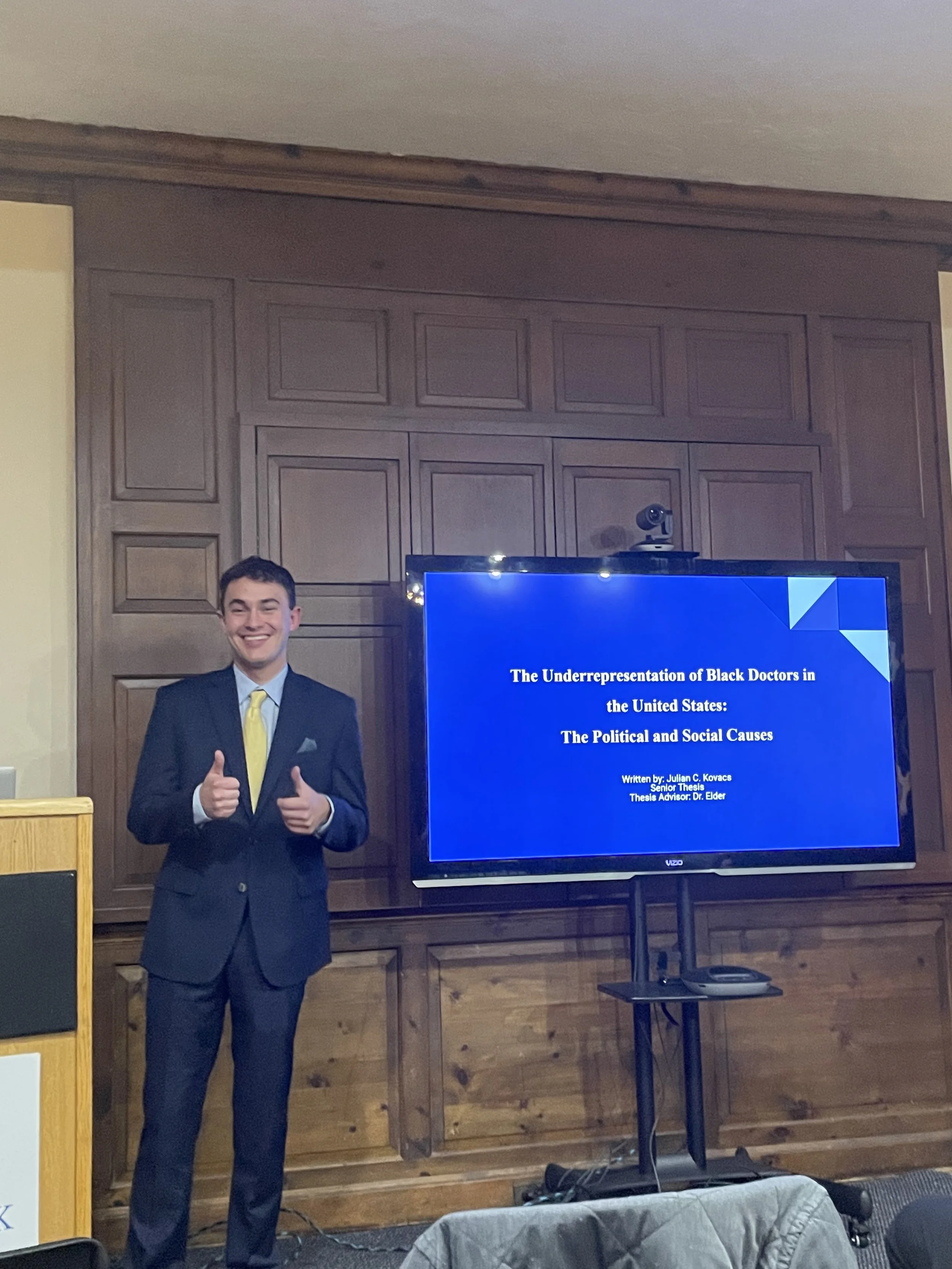 A young man in a suit and yellow tie giving a thumbs-up next to a presentation slide on a television screen in a wood-paneled room.