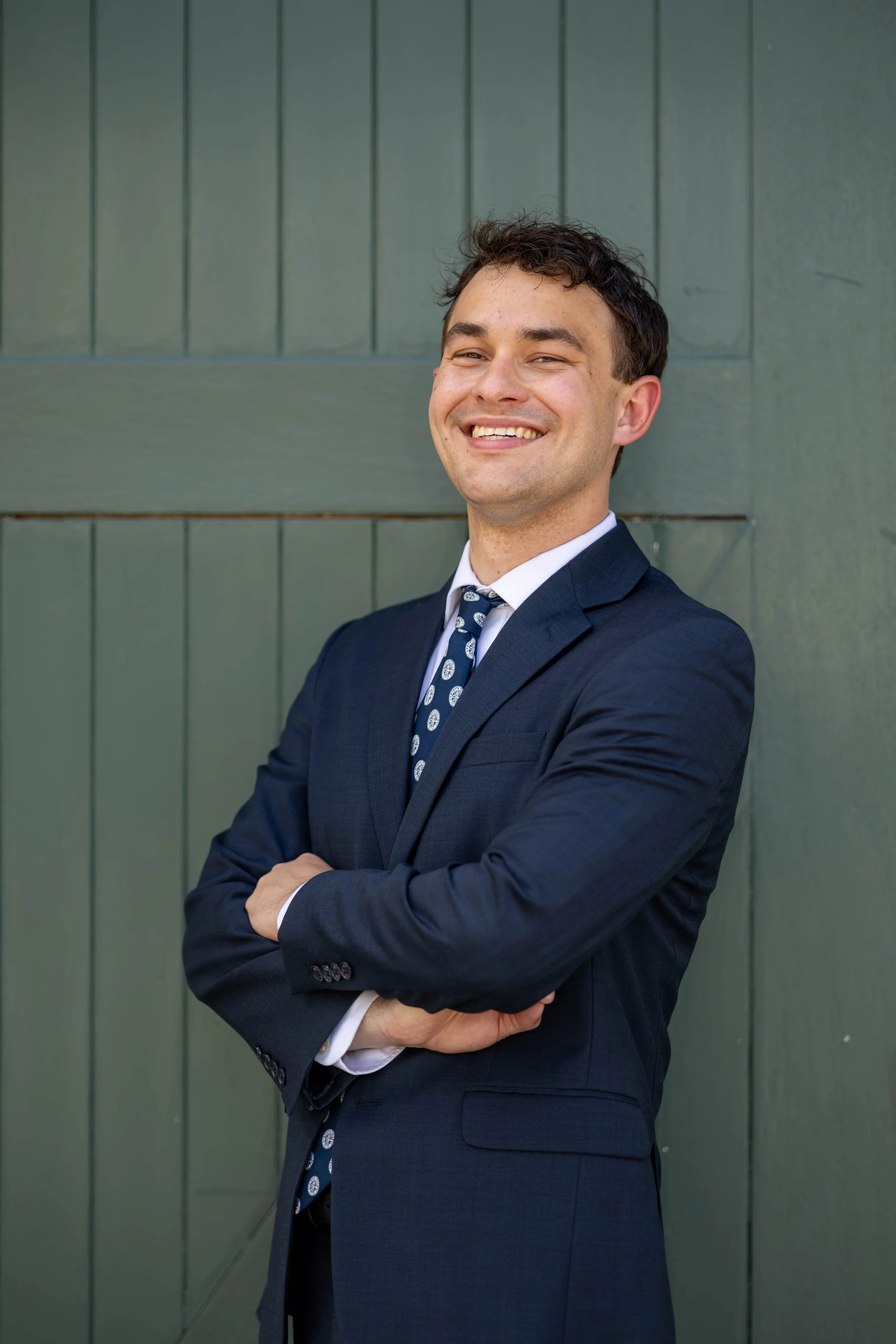 A young man in a navy suit with a white shirt and patterned tie, smiling with arms crossed, standing against a green wooden wall.