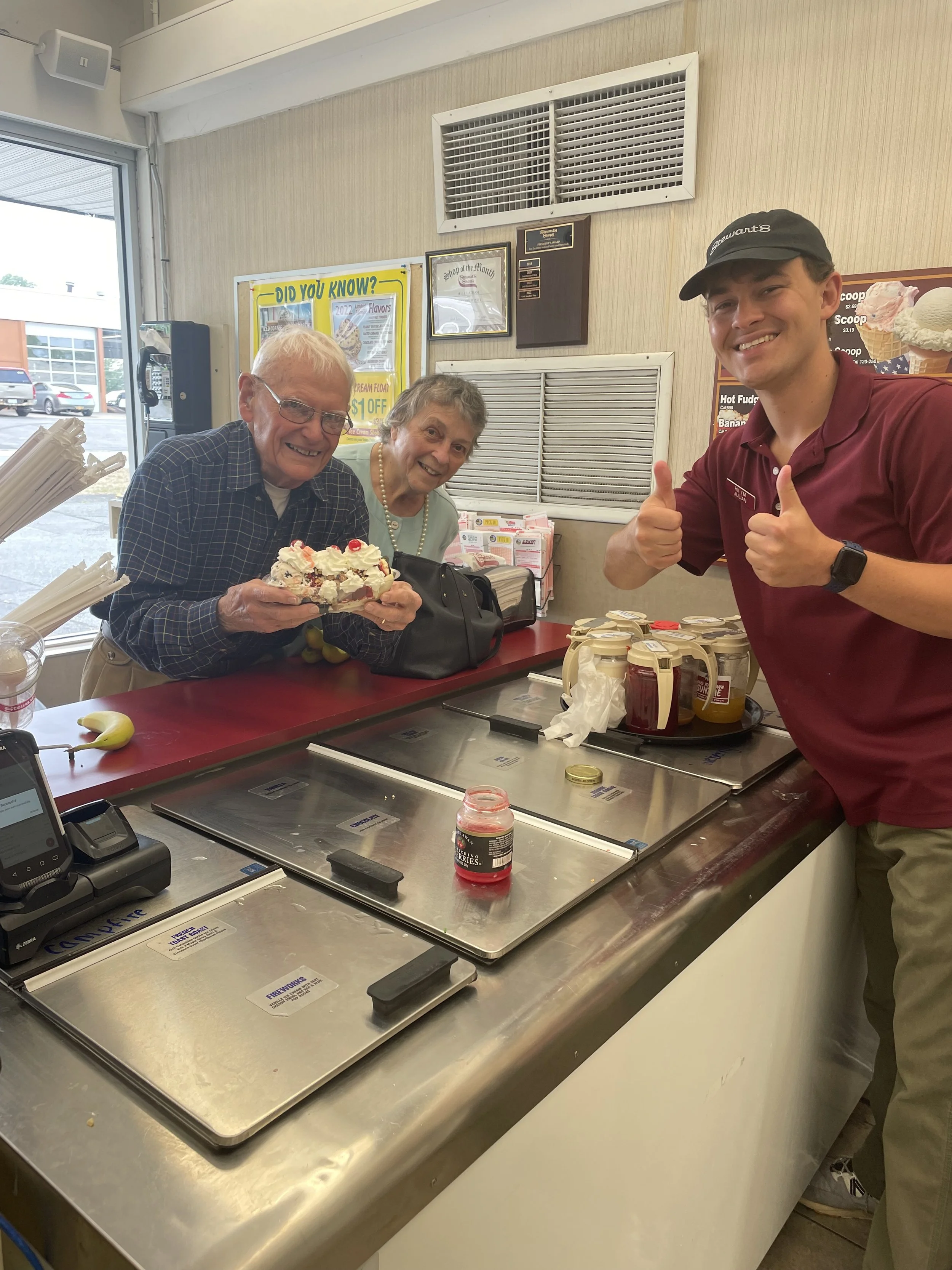 An elderly man and woman smiling and holding a cake inside an ice cream shop, with a young male employee giving two thumbs-up.