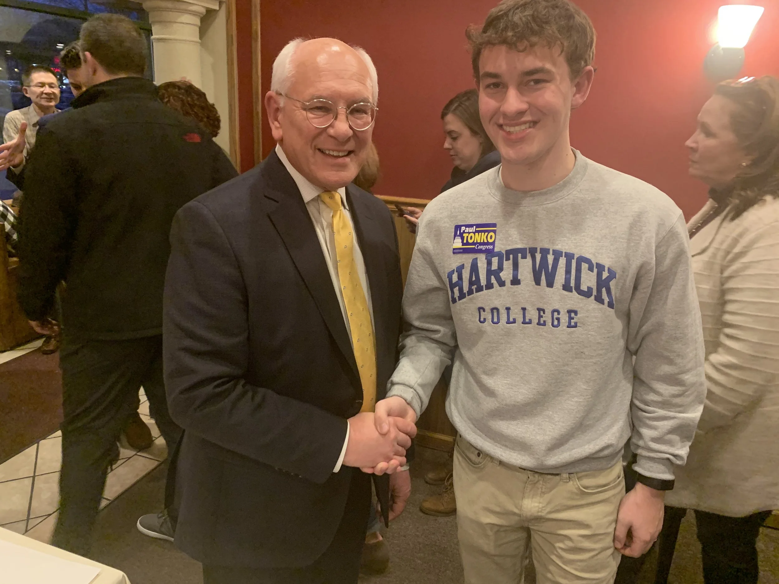 Two men shaking hands at an indoor social event; one is an older man in a dark suit with a yellow tie and glasses, the other is a young man in a gray sweatshirt with "Hartwick College" written on it, wearing a Abe Lincoln campaign sticker.