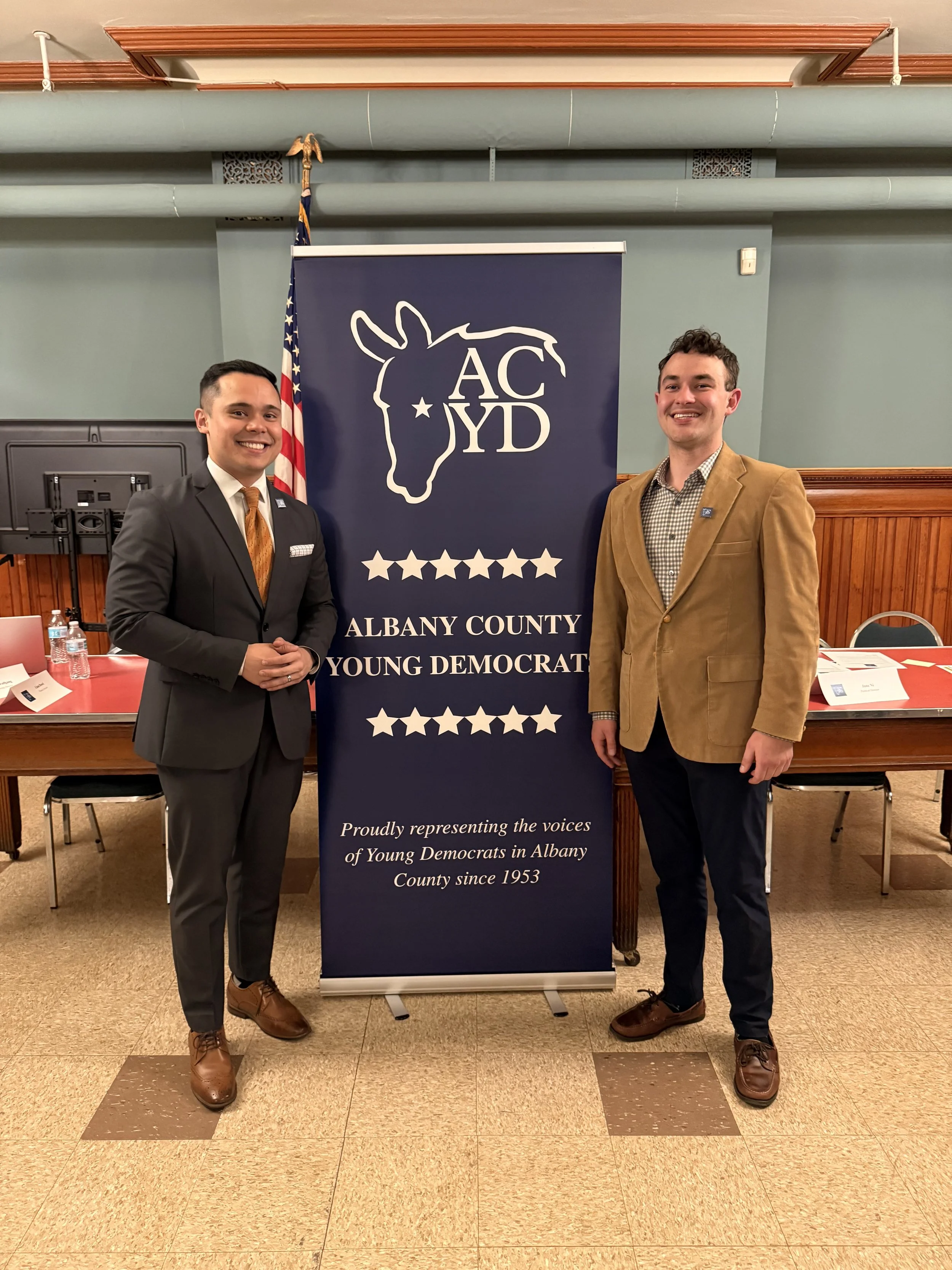 Two young men in suits standing on either side of a large blue banner for the Albany County Young Democrats, in a room with an American flag and tables with documents. One man wears a dark suit with a gold tie, the other a brown blazer with a checkered shirt.
