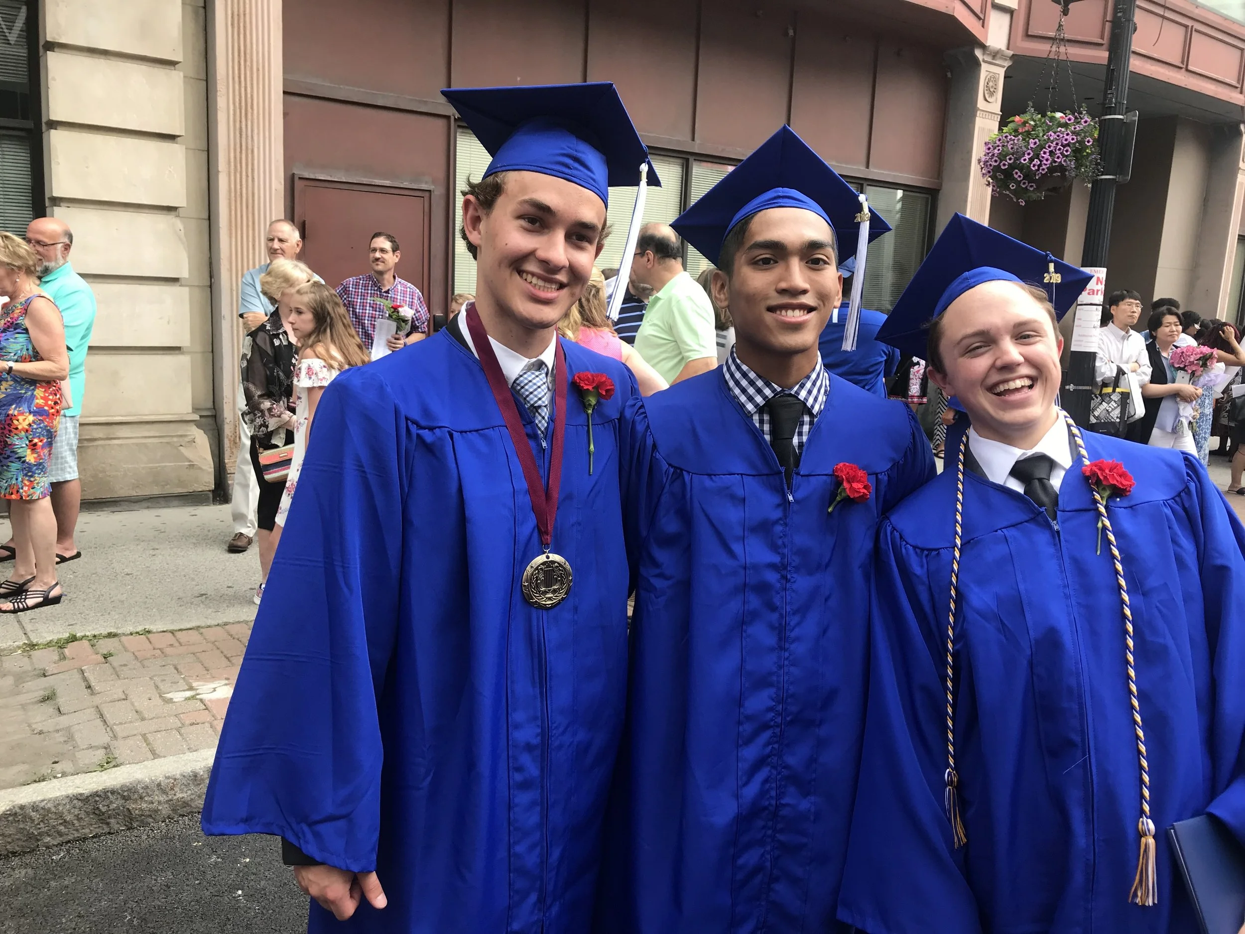 Three graduates wearing blue caps and gowns, with red carnations pinned to their gowns, smiling for a photo during a graduation ceremony outdoors.