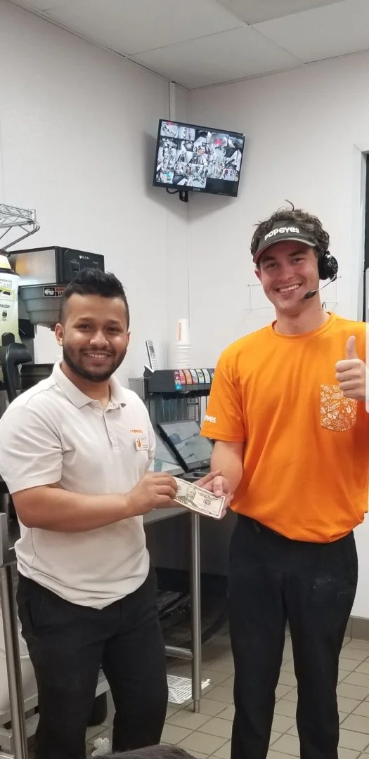 Two employees in a fast-food restaurant exchanging money, one smiling and giving a thumbs-up, with a TV screen displaying security camera footage above them.