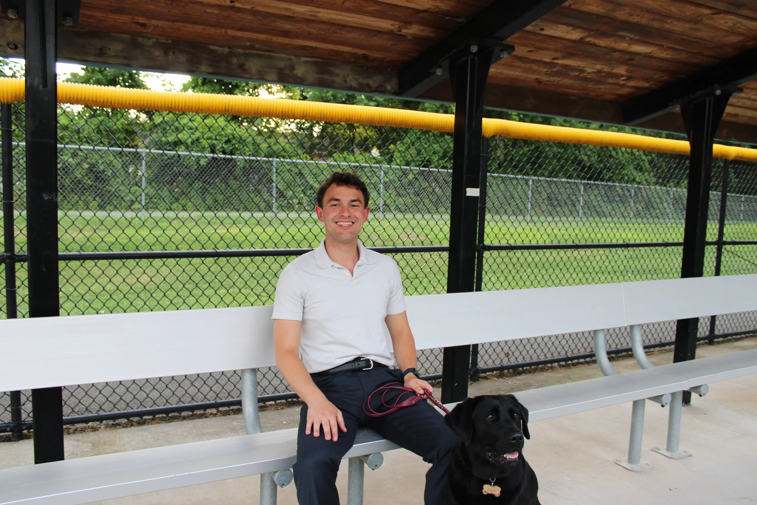 A young man in a white polo shirt and dark pants sitting on a silver and white park bench with a black Labrador Retriever dog on his lap, under a wooden shelter at a park with a chain-link fence and green grass in the background.