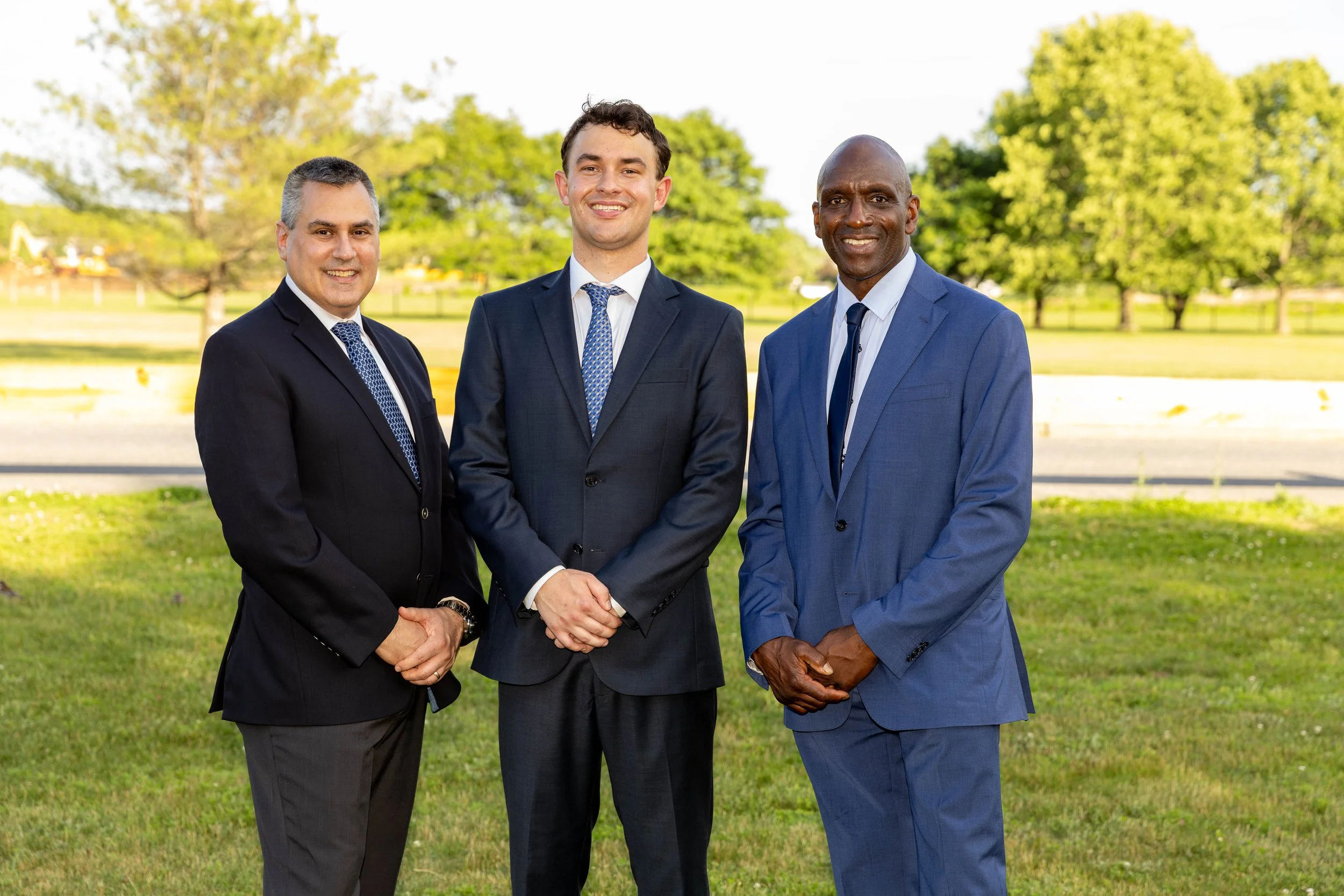 Three men in suits standing outdoors on a grassy area with trees and a road in the background, smiling at the camera.