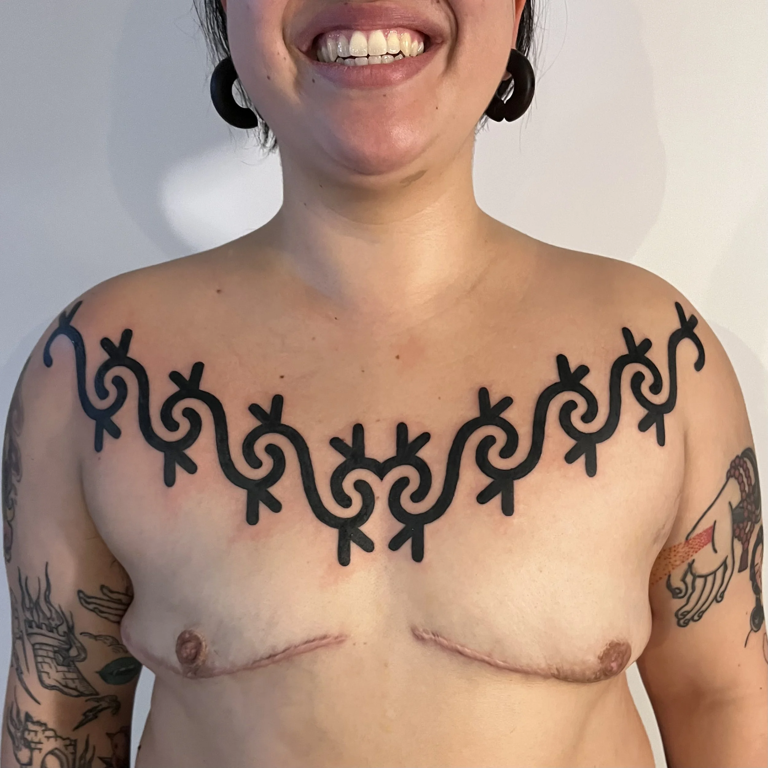 A person smiling and showing their upper body, with tattoos on the arms and chest, and a large black tattoo across the upper chest.