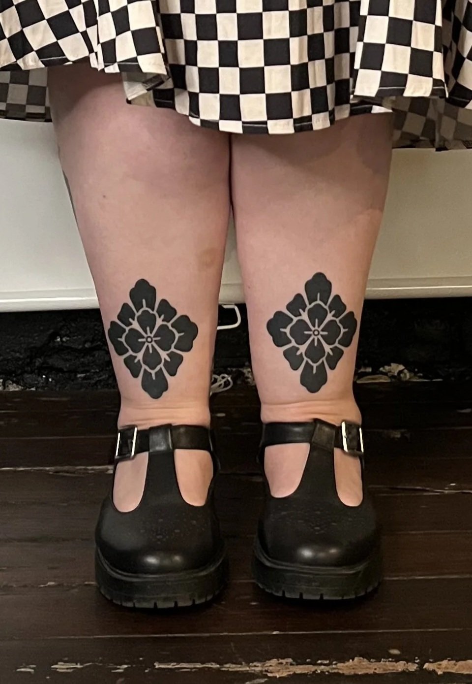 Person wearing black shoes with matching floral tattoos on ankles, a checkered skirt, and a quilted brown jacket, standing on a dark wooden floor against a white wall.