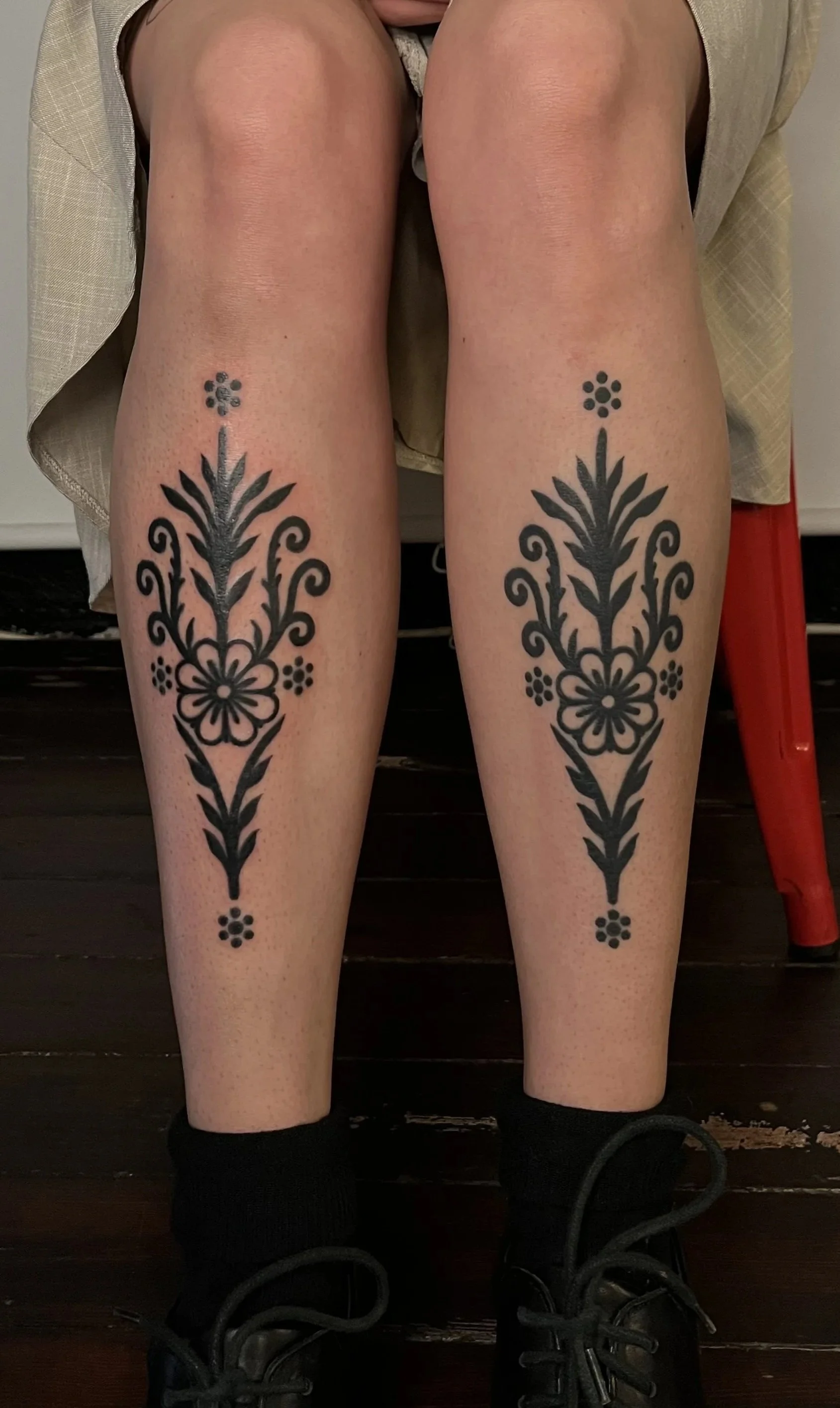 Two person's lower legs with black floral tattoos, wearing black shoes and socks, seated on a dark wooden floor.