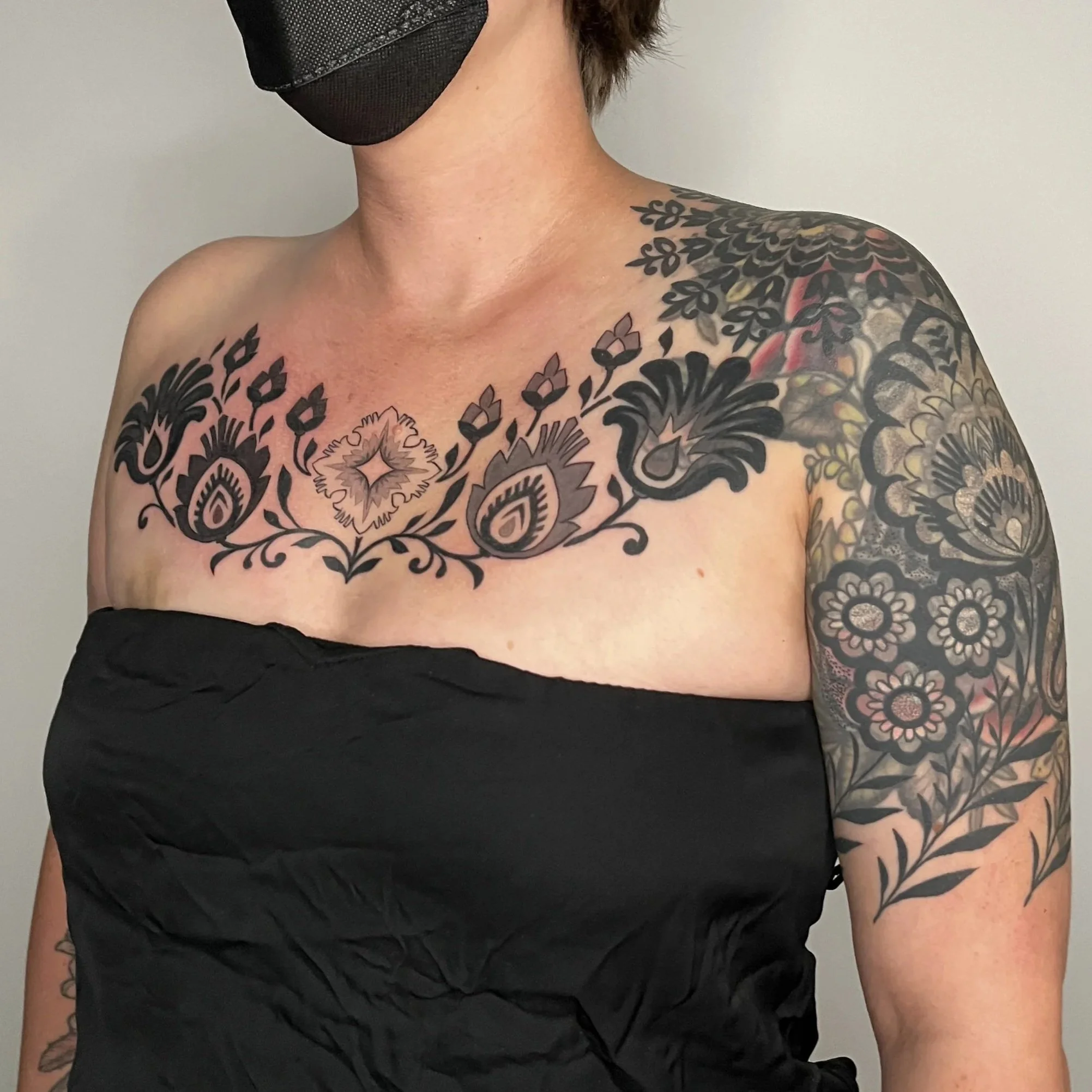A person with short brown hair, wearing glasses and a black face mask, stands against a plain wall. They are showing several large, colorful tattoos on her chest, shoulders, and arms. 