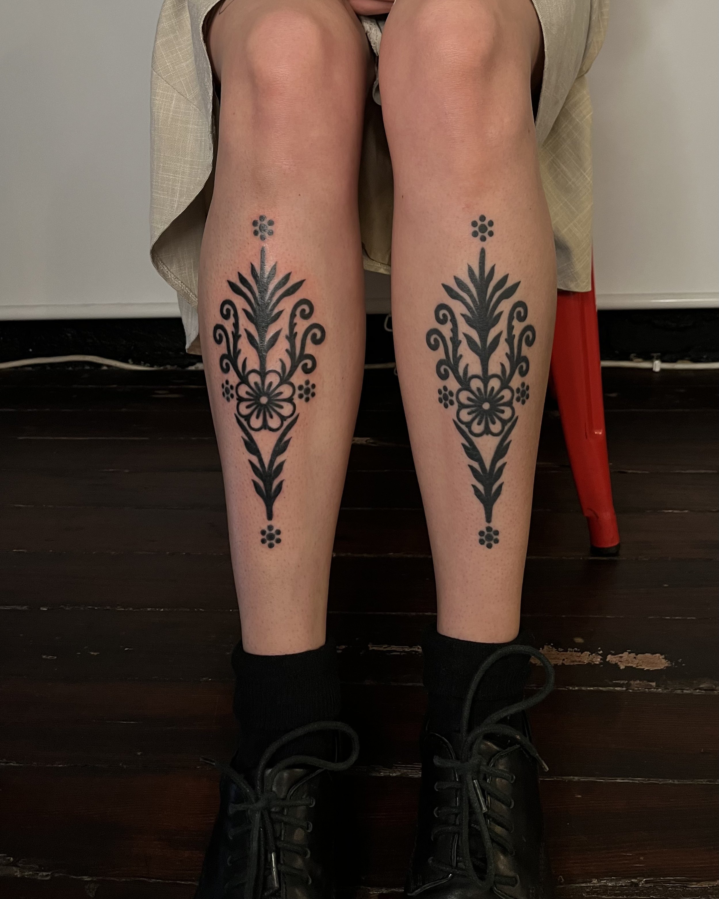 Two person's lower legs with black floral tattoos, wearing black shoes and socks, seated on a dark wooden floor.