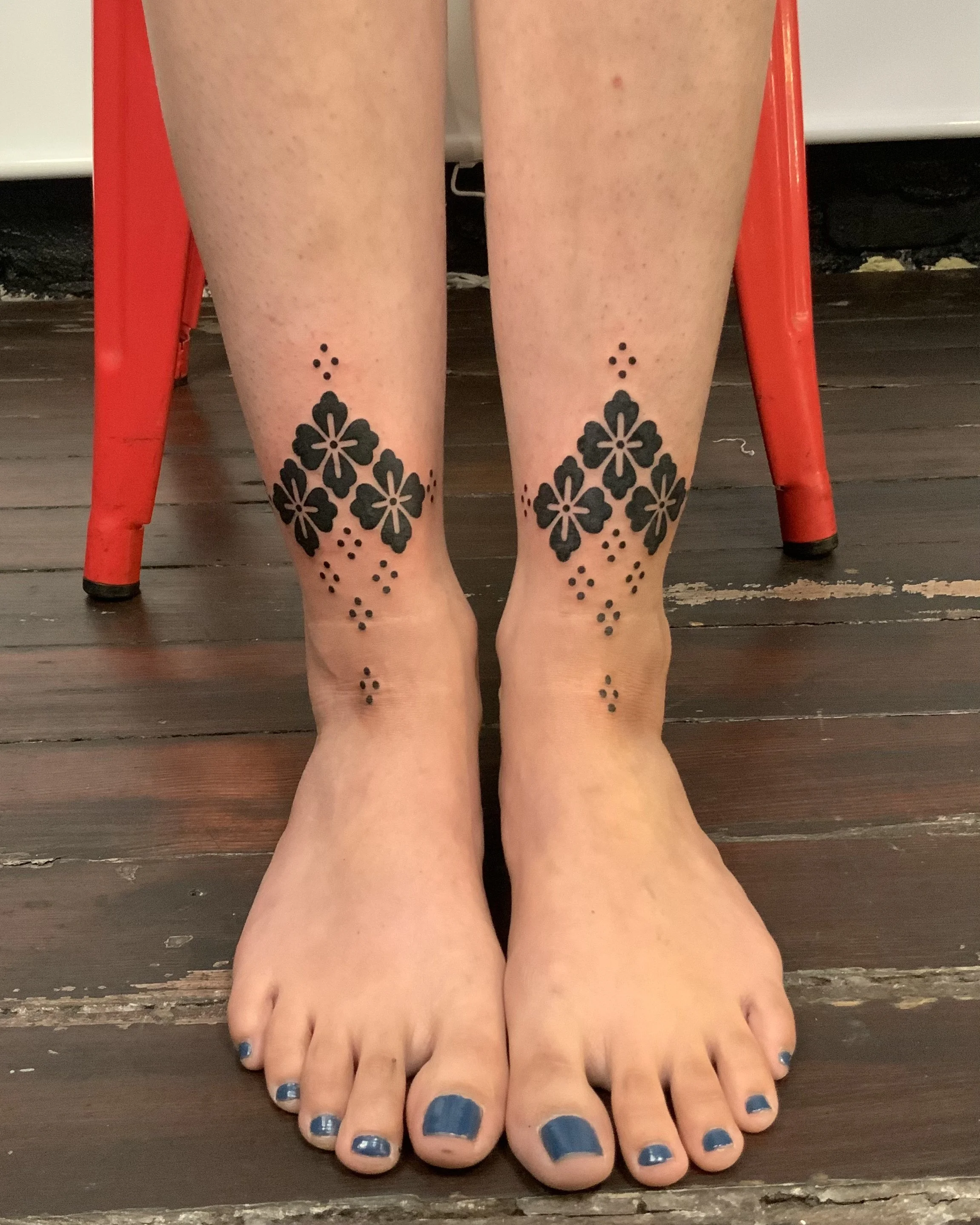 Person sitting on a chair showing their feet with a black floral tattoo on the lower legs and blue painted toenails.