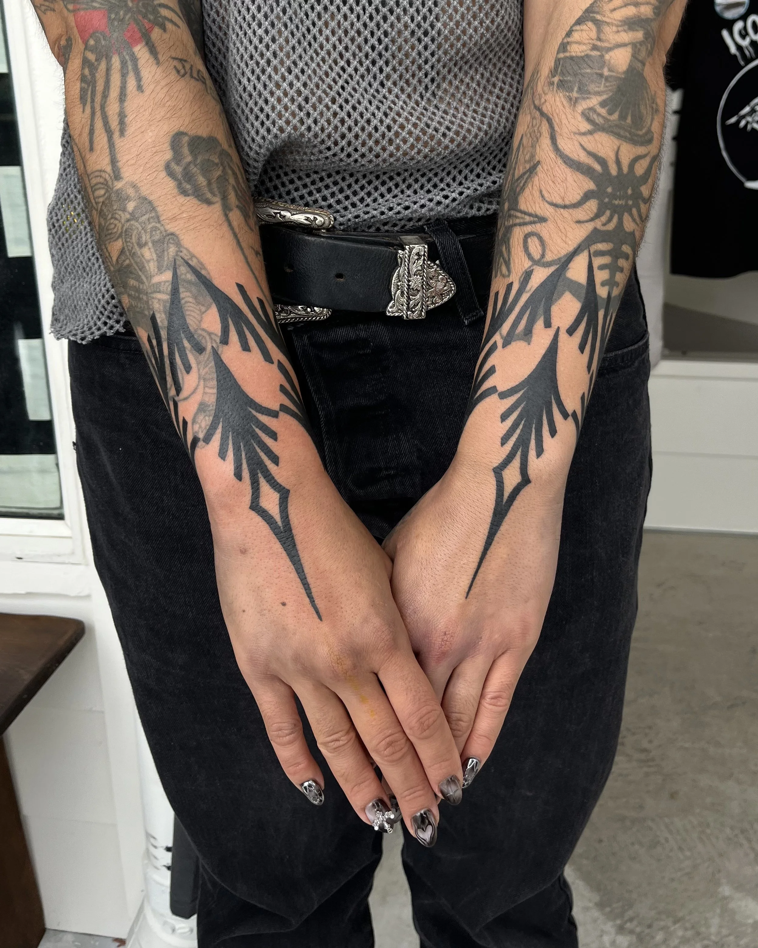 Person with tattooed arms wearing a mesh shirt, black jeans, and a belt, with hands clasped in front.