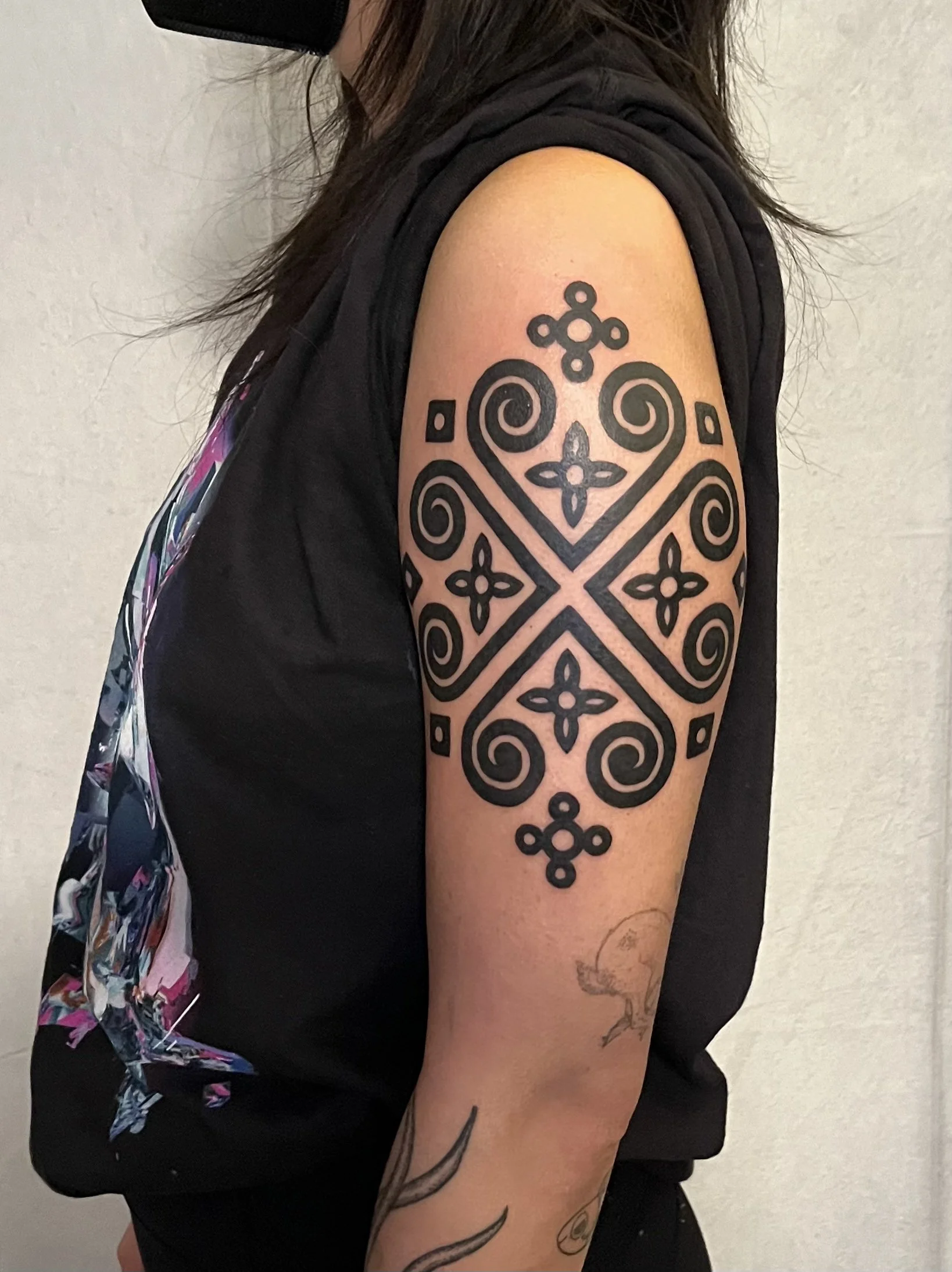A person's upper arm with a large, intricate black tribal-style tattoo featuring symmetrical swirls, flowers, and geometric patterns.