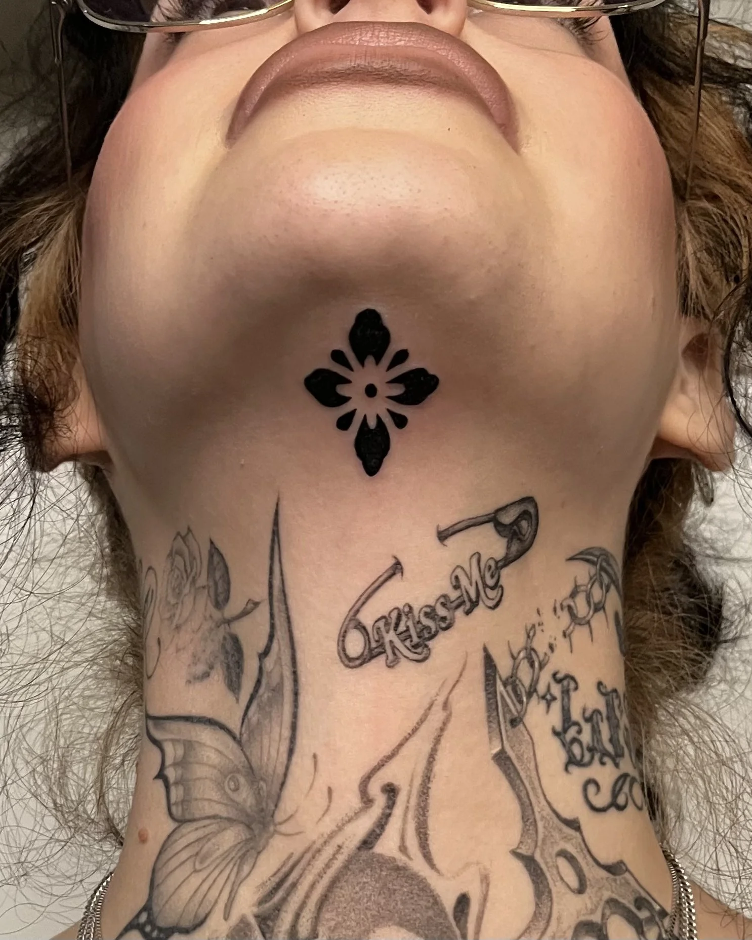 Close-up of a person's chin and neck with various tattoos, including a black floral design above the chin.