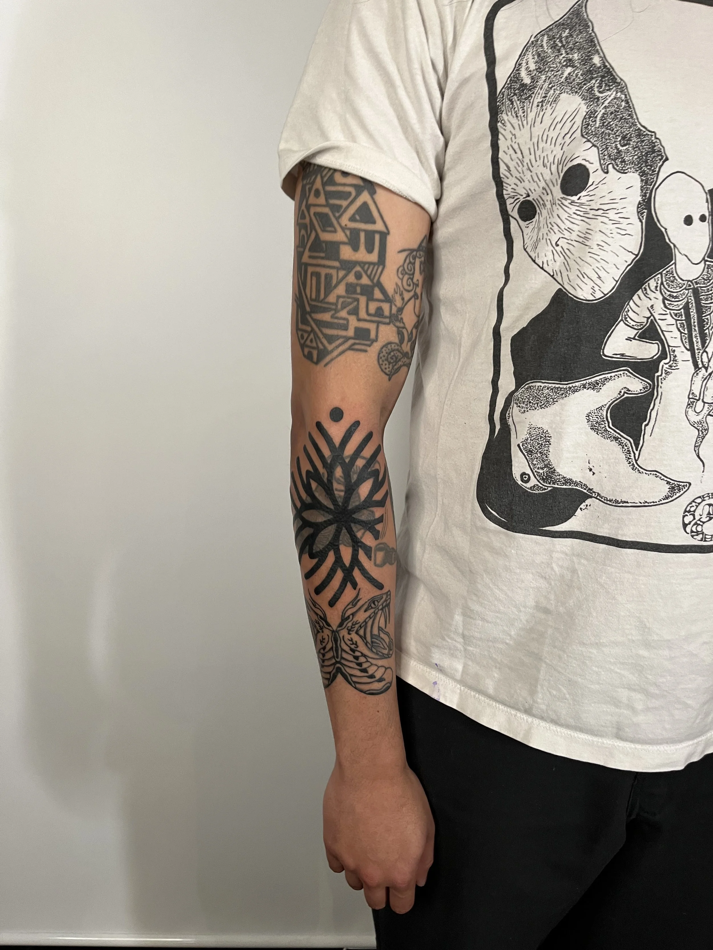 Person's arm with tattoos including a black tribal design, a snowflake, a moth, and a snake, wearing a white T-shirt with graphic black and white illustrations.
