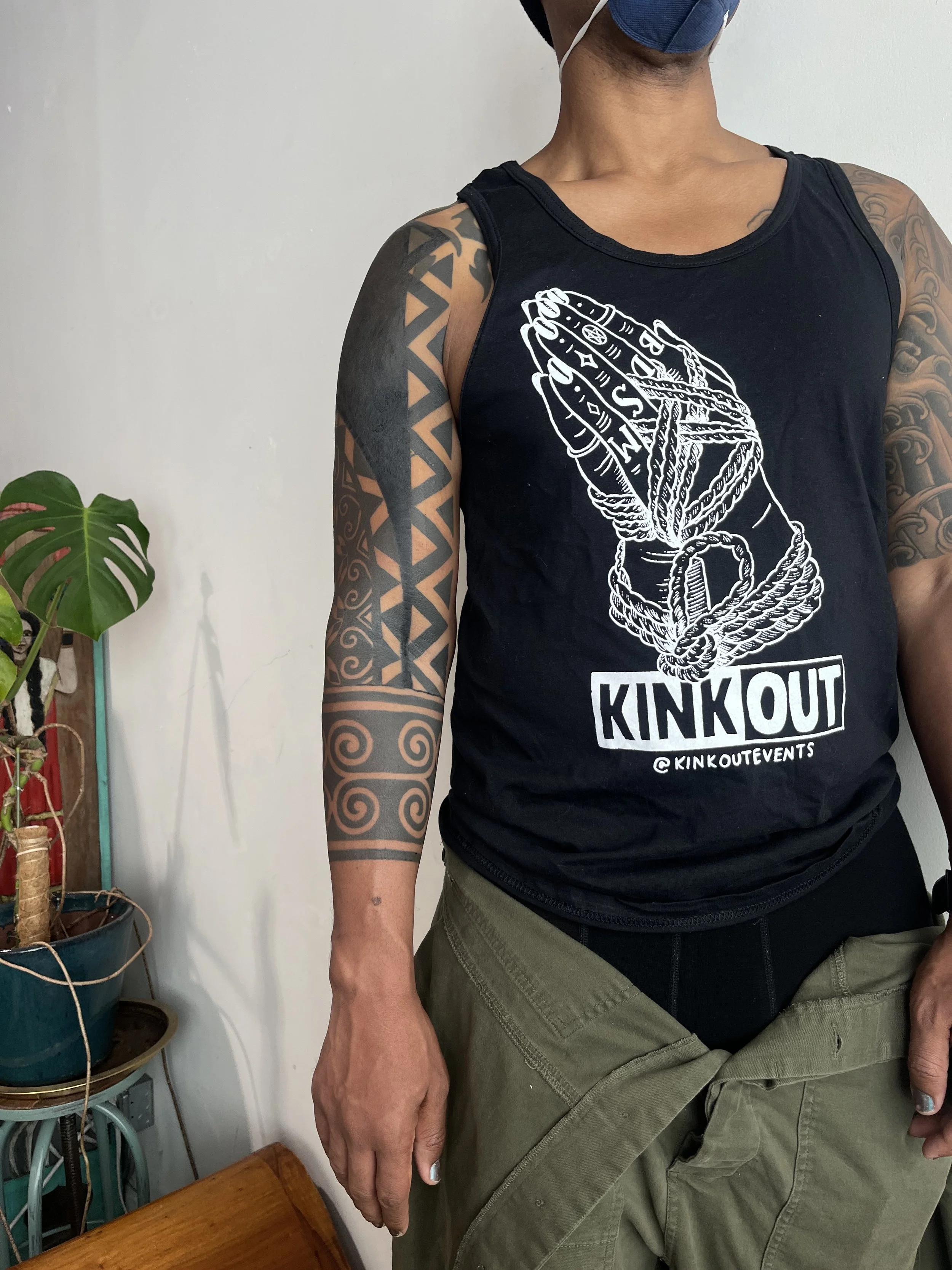 A person with tattoos on their arm wearing a sleeveless black T-shirt with white graphic and text, khaki cargo pants, and a blue face mask, standing indoors near a potted plant.