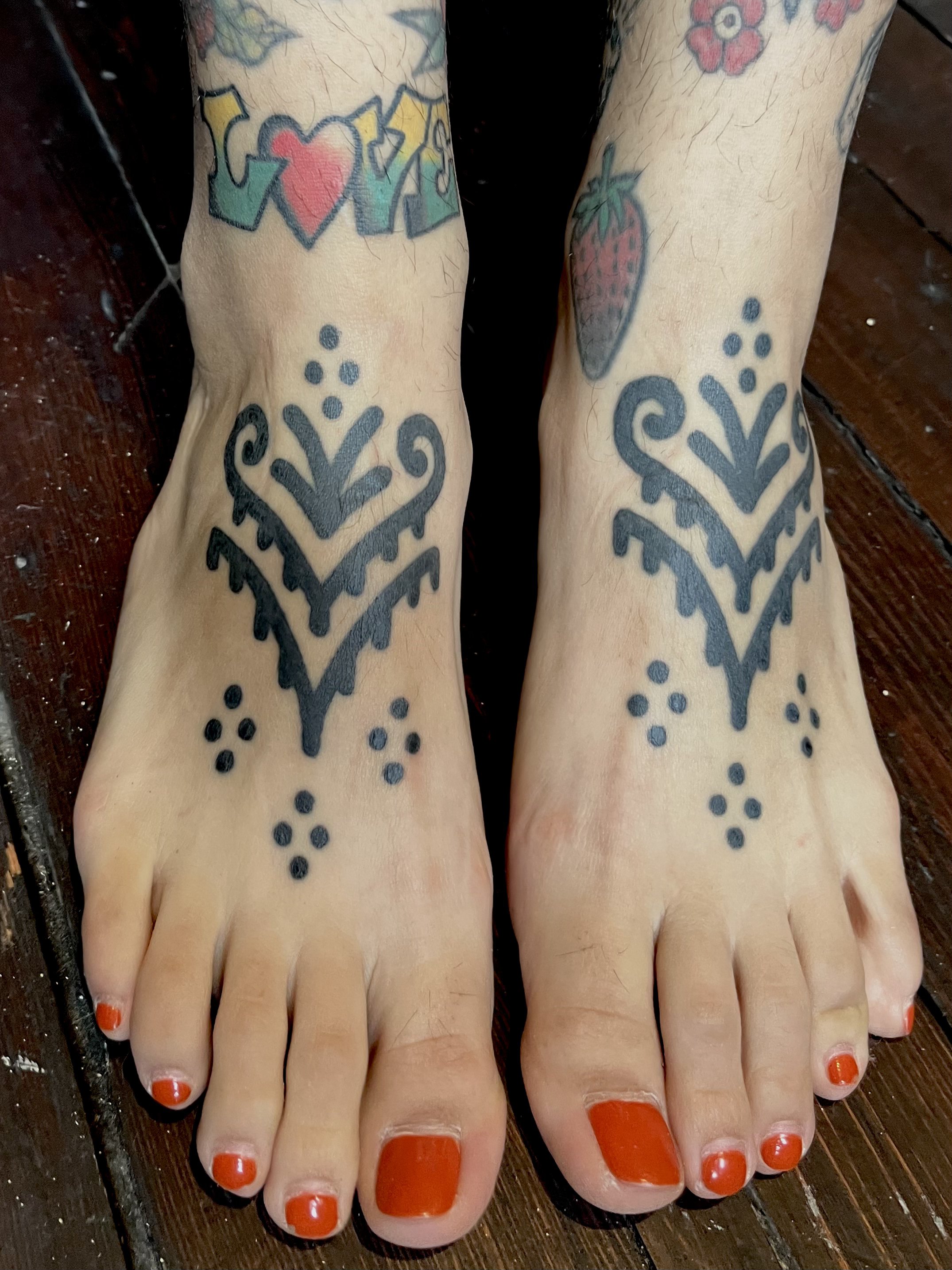 Tattooed feet with black tribal designs on the top of each foot and red painted toenails, placed on a wooden floor.