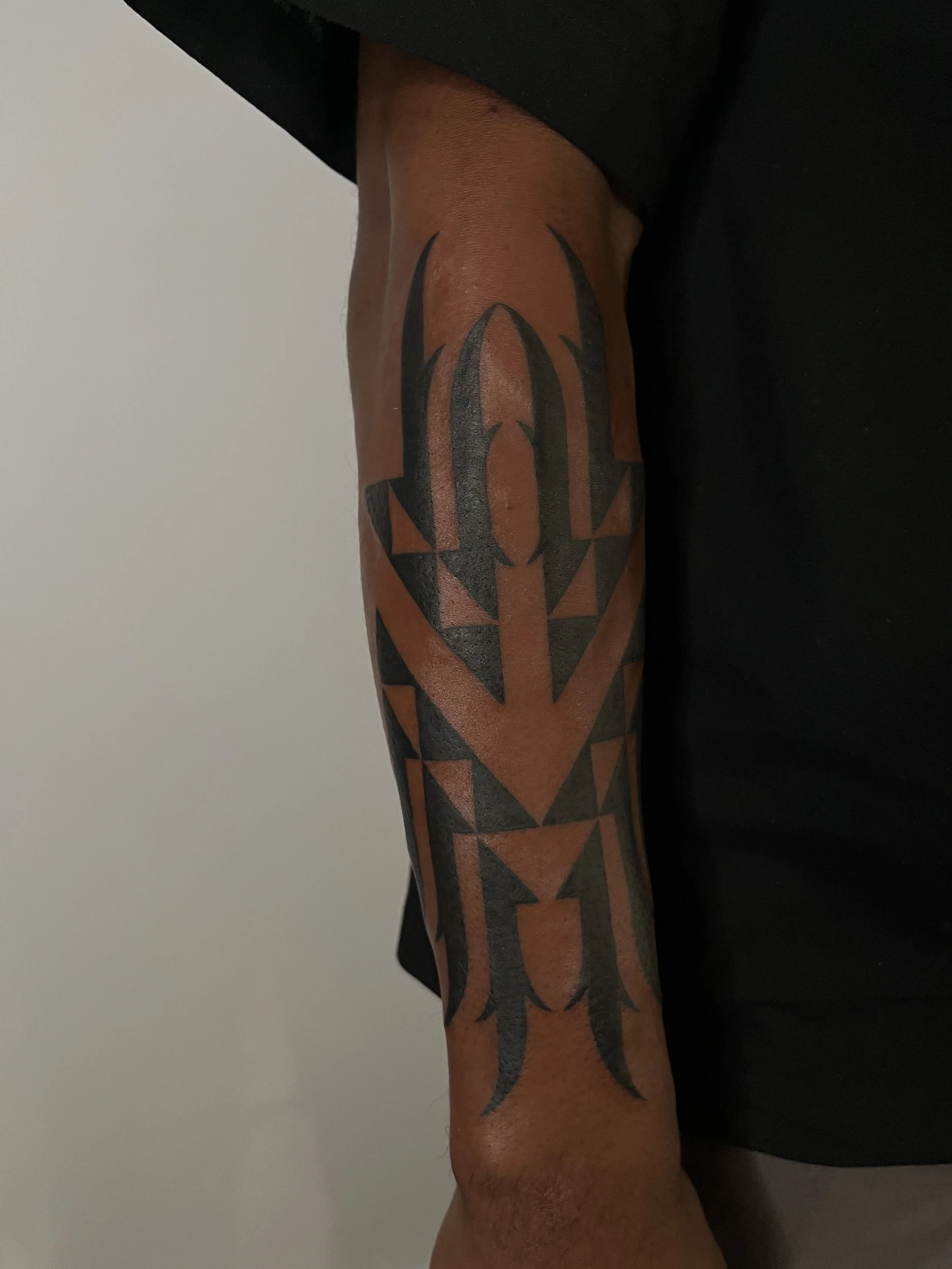 A person's upper arm with a black tribal tattoo featuring sharp pointed shapes and symmetrical patterns.