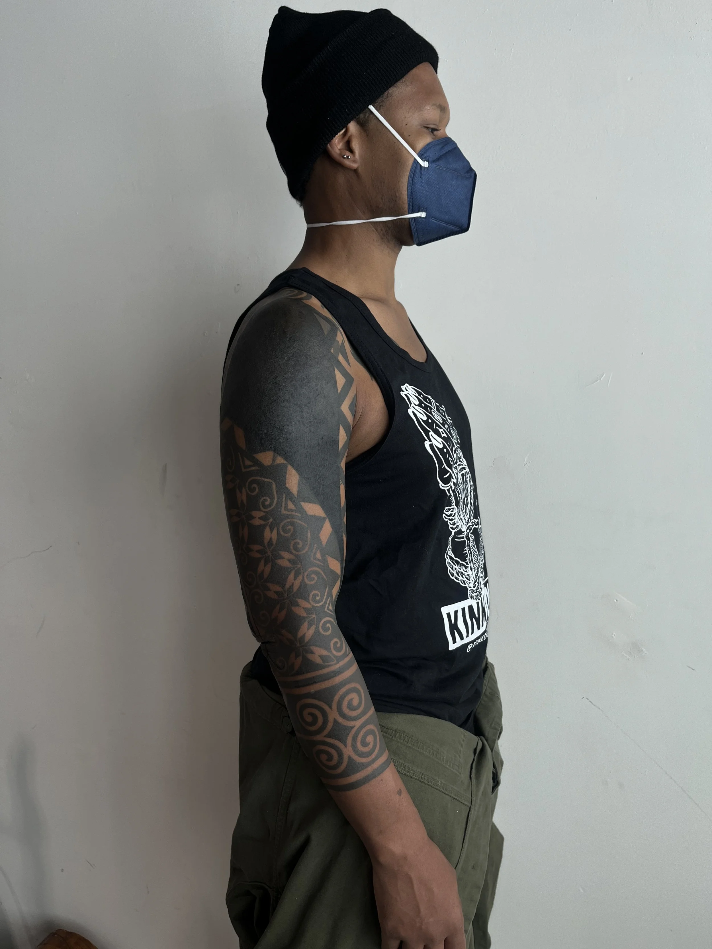A person standing sideways wearing a black beanie, a dark blue face mask, a sleeveless black graphic T-shirt, and olive green cargo pants, with tattoos on their left arm.