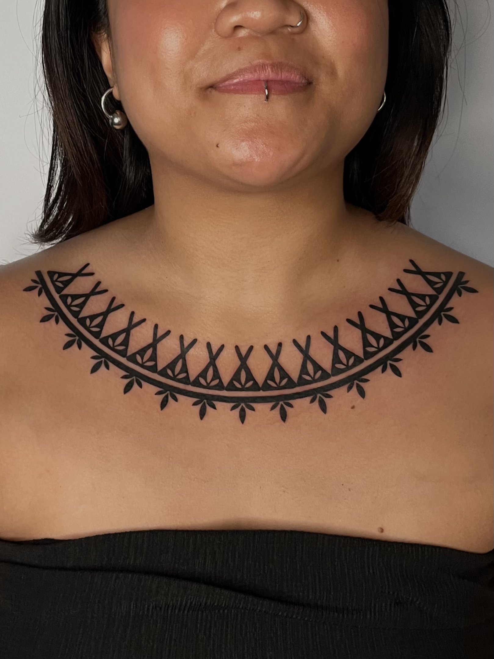 A person displays a black tattoo around their shoulders that resembles a decorative necklace pattern with geometric shapes.