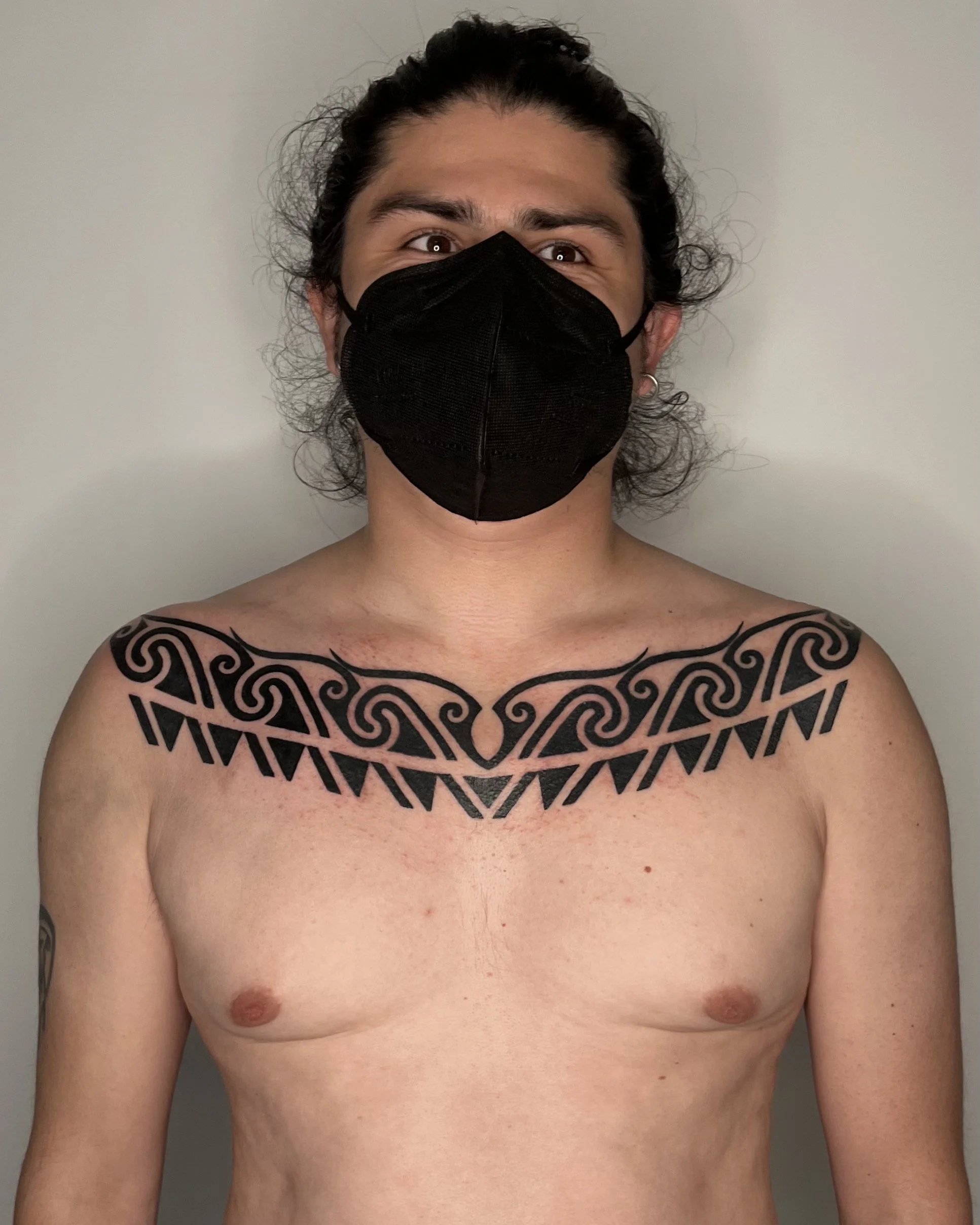 A person with long curly hair, wearing a black face mask, and a black tribal-style tattoo across the chest, standing shirtless against a plain gray background.
