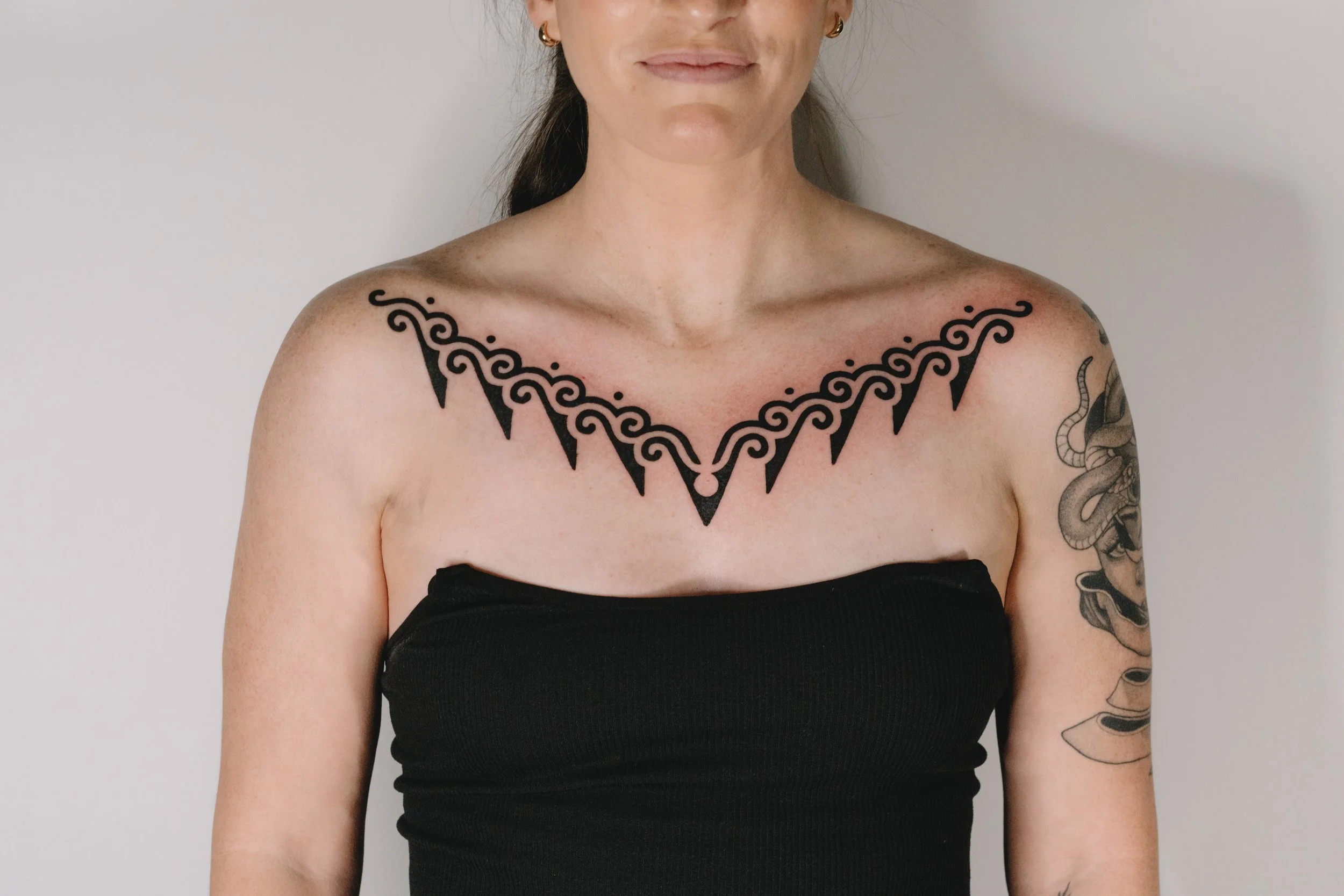 A person with black tribal tattoo across shoulders and upper chest, wearing a strapless black top against a plain light-colored wall.