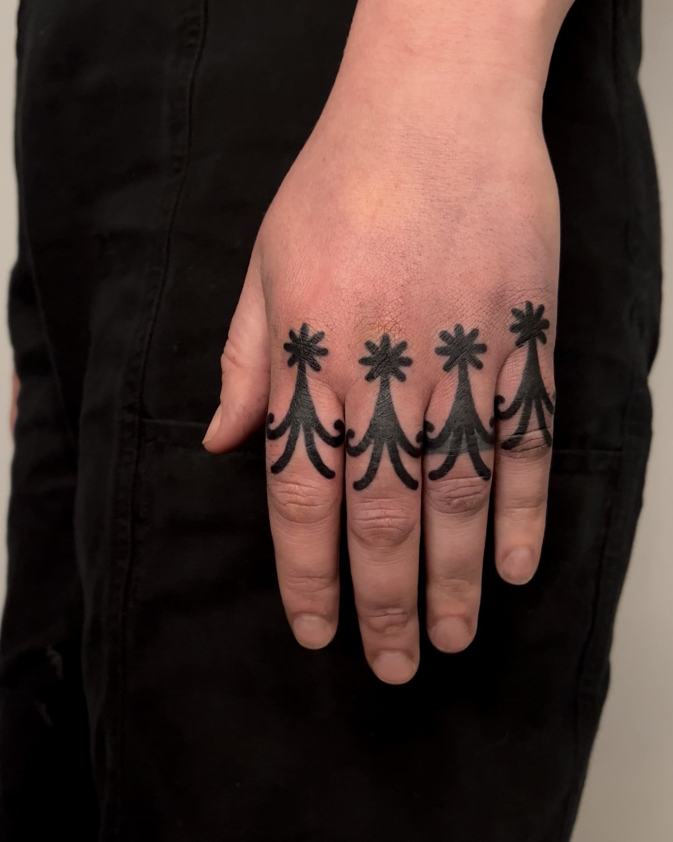A person's hand with a tattoo of black flower-like designs across the fingers, standing against a background of black pants.