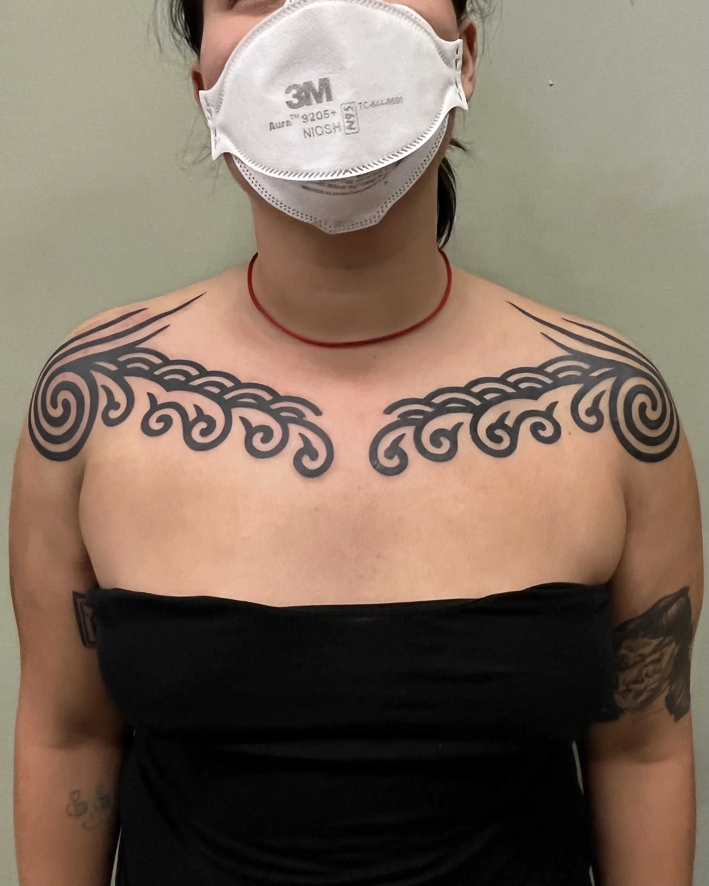 Person with shoulder tattoos wearing a black strapless top, red choker, and a white N95 mask standing against a plain wall.