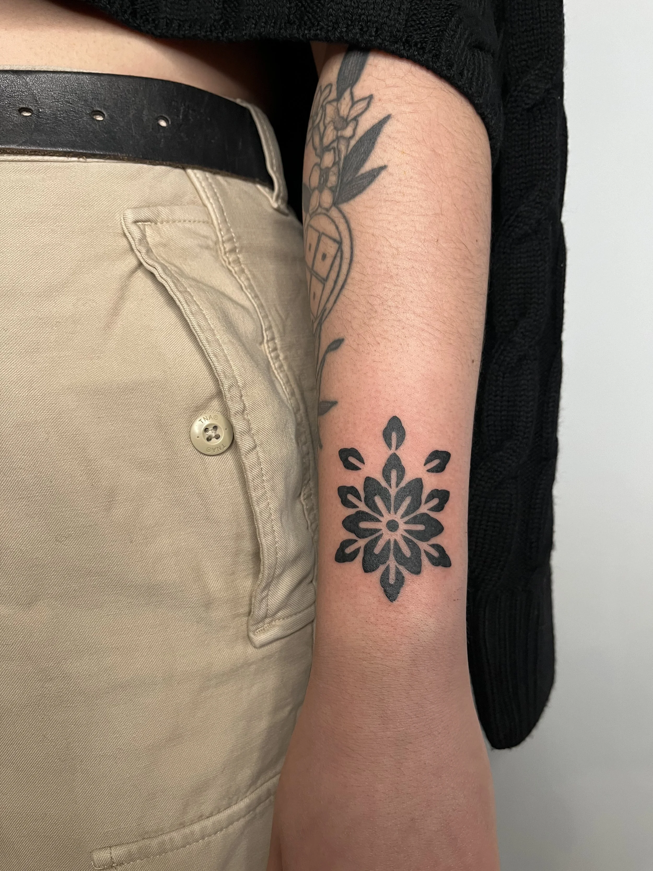 Person's arm with a black floral tattoo, wearing beige pants and a black belt, with part of a tattooed sleeve visible.
