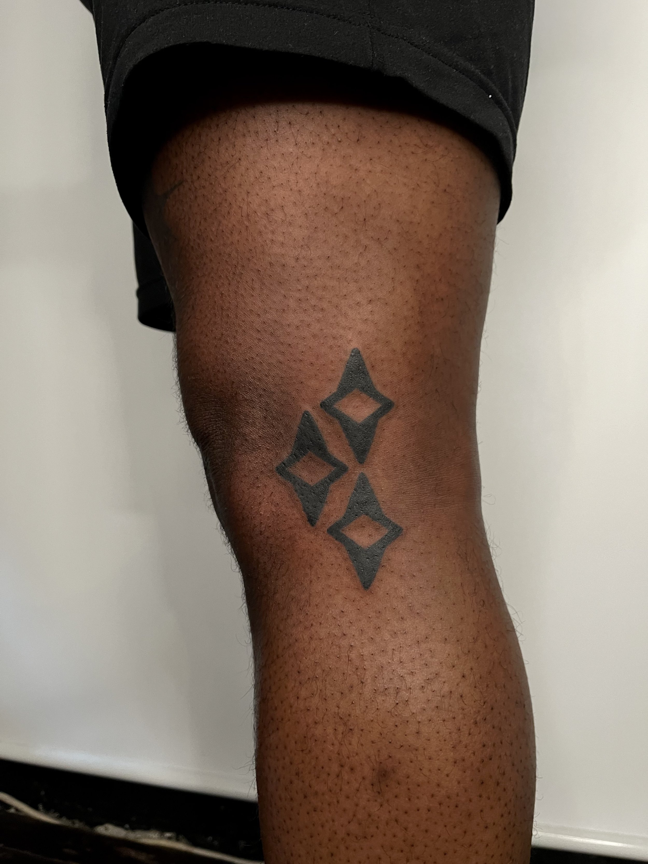 A person's leg wearing black shorts, showing a black tattoo of four diamonds arranged vertically on the lower thigh.