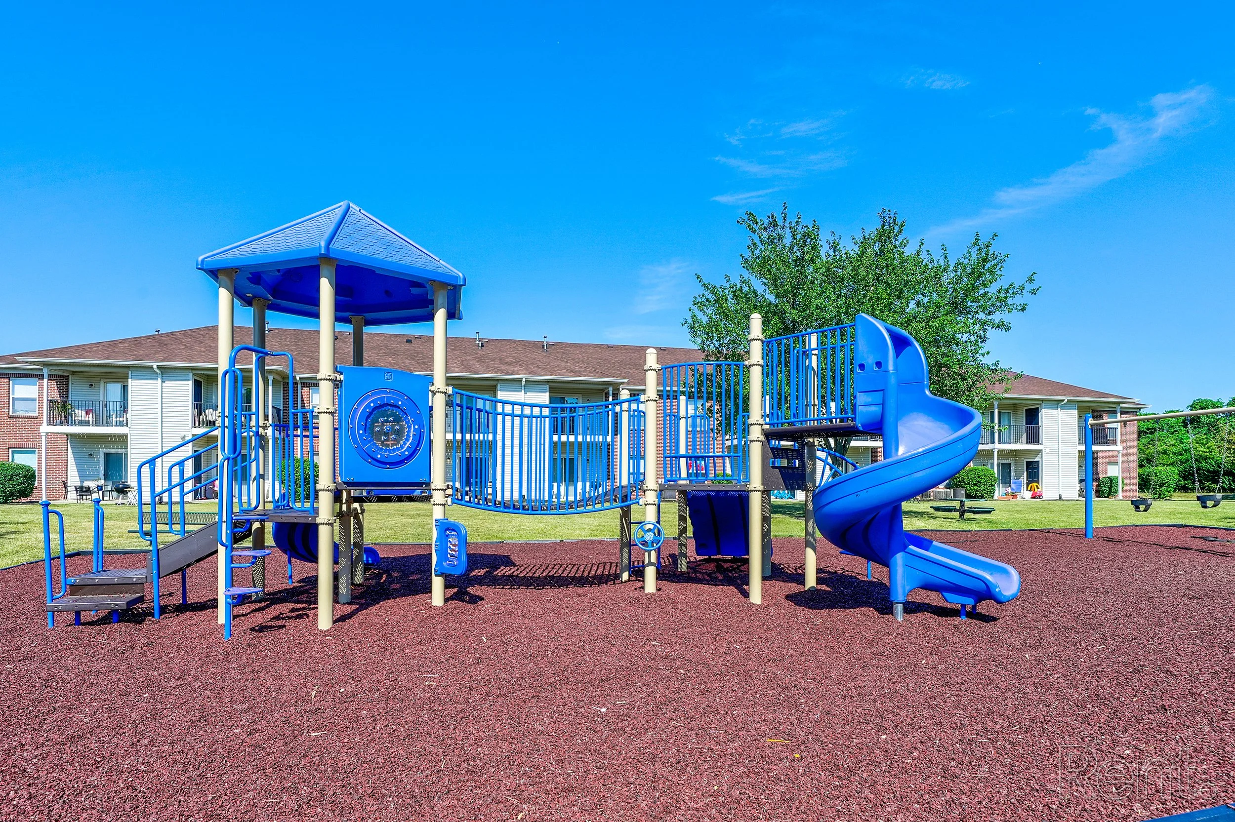 Blue playground structure with slides, bridges, and climbing elements on red rubber surfacing, residential buildings in background, clear blue sky.