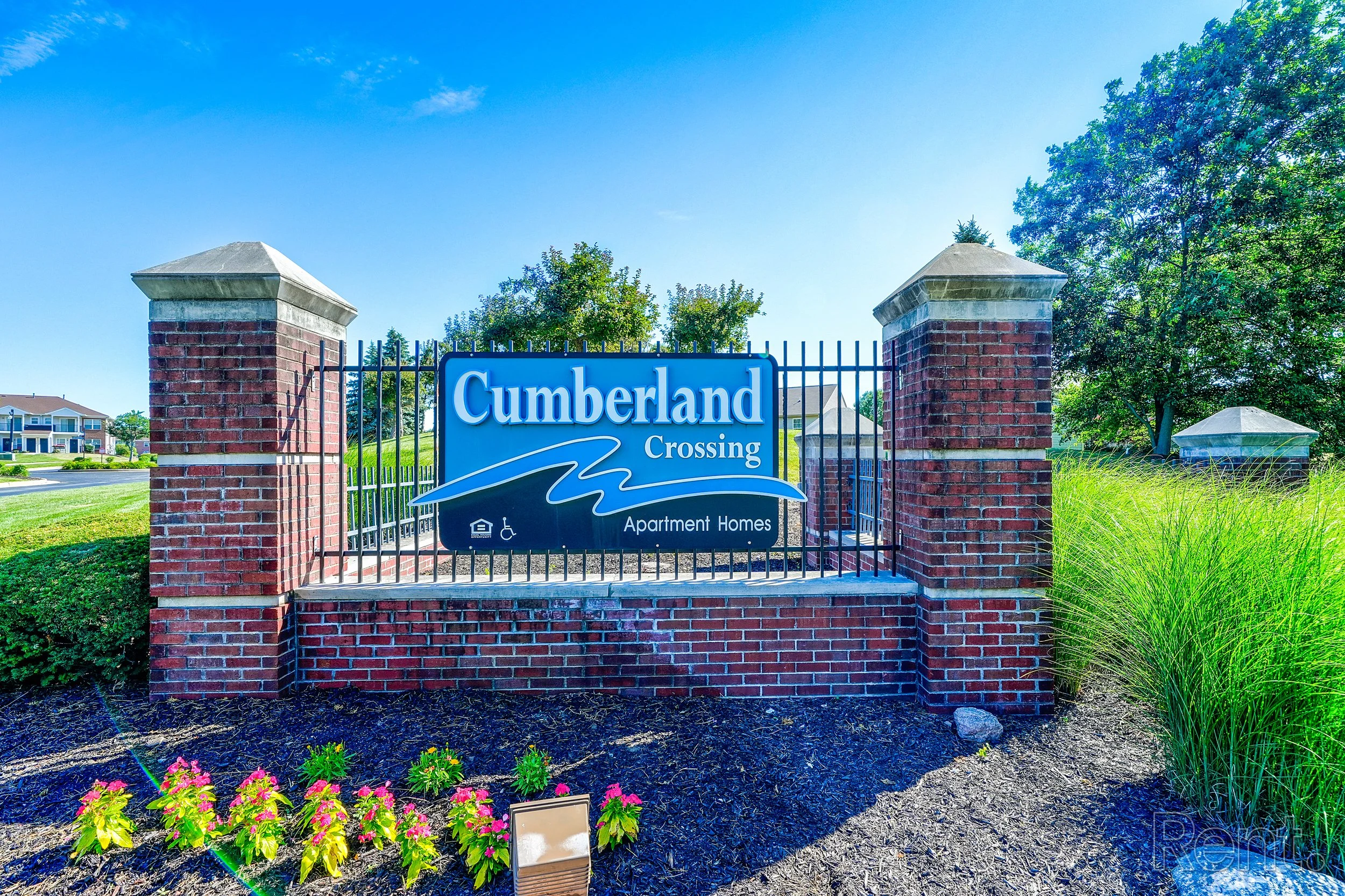 Entrance sign for Cumberland Crossing Apartment Homes with brick pillars and metal gate, surrounded by landscaping and greenery.