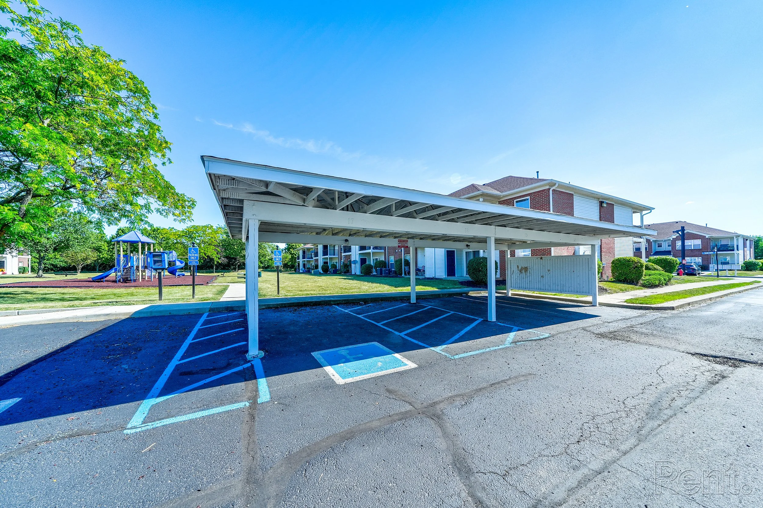 Empty parking lot with a covered parking space, residential buildings, a playground, trees, and signs indicating accessible parking, under a clear blue sky.