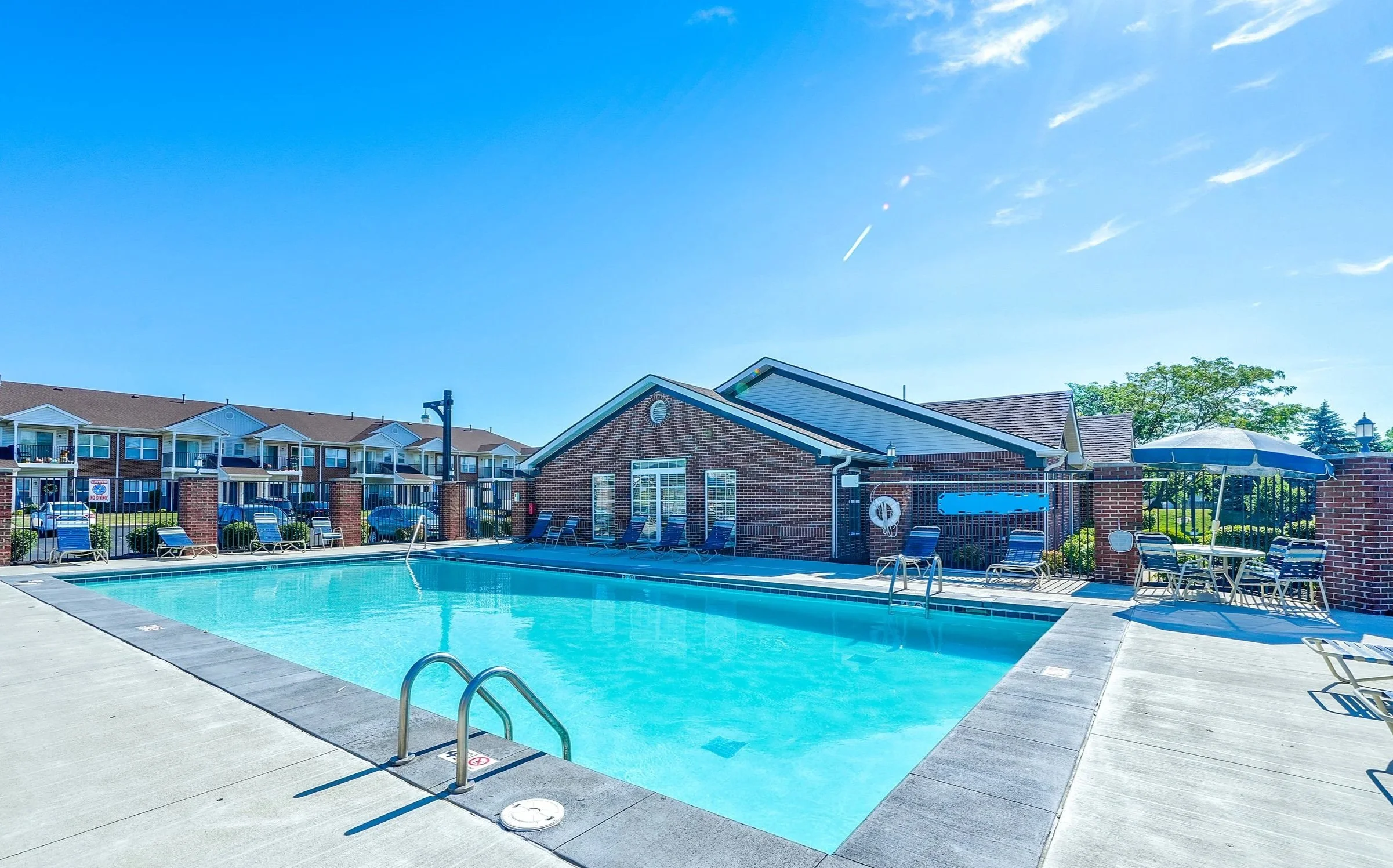 An outdoor swimming pool in Fishers with blue water, surrounded by lounge chairs, tables with umbrellas, and a brick building. In the background are apartment complex buildings under a clear blue sky.