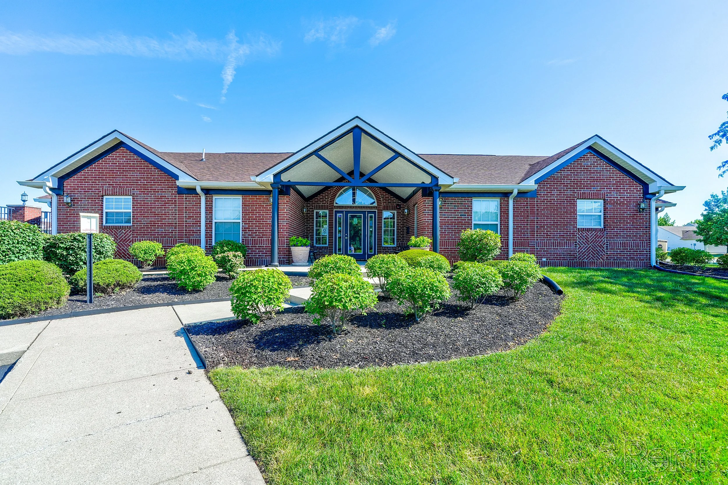 A brick Leasing Office surrounded by green bushes and grass, under a clear blue sky.