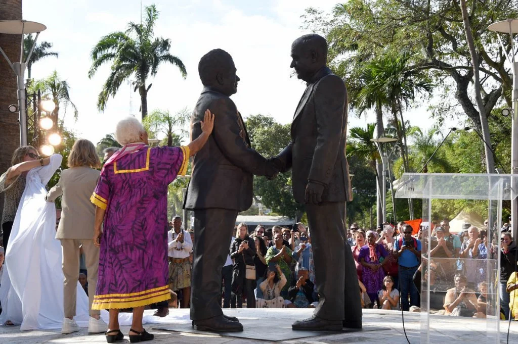 Deux hommes en costume se serrent la main lors d'une cérémonie en plein air, entourés de personnes et de palmiers.