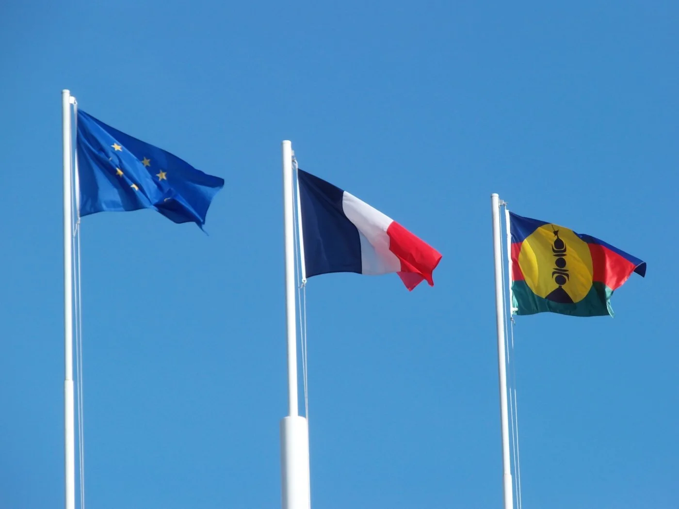 Drapeaux de l'Union européenne, de la France et du FLNKS flottant sous un ciel bleu