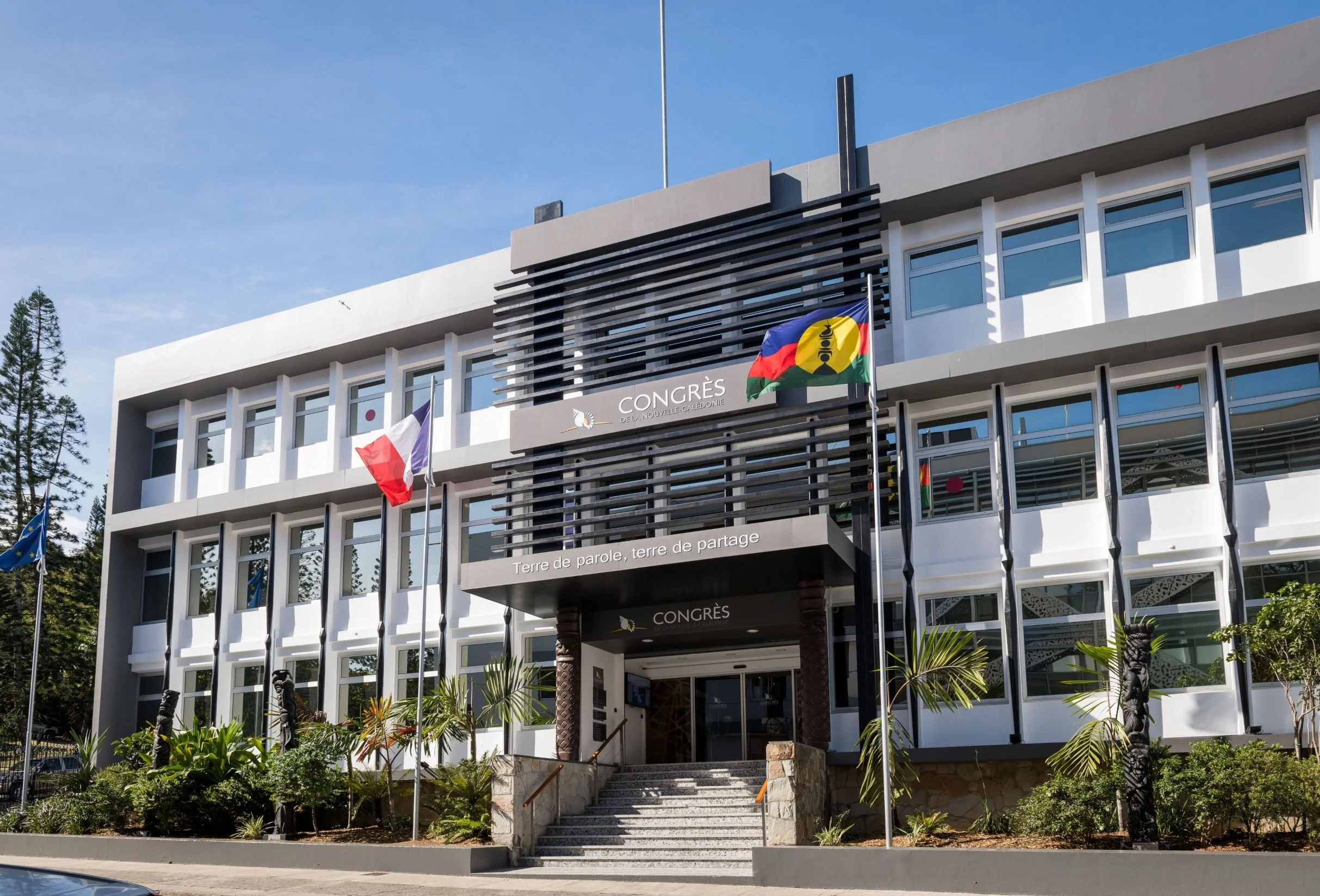 Bâtiment moderne avec drapeaux français, calédonien et européen flotant devant le Congrès de Nouvelle-Calédonie, avec végétation tropicale autour.