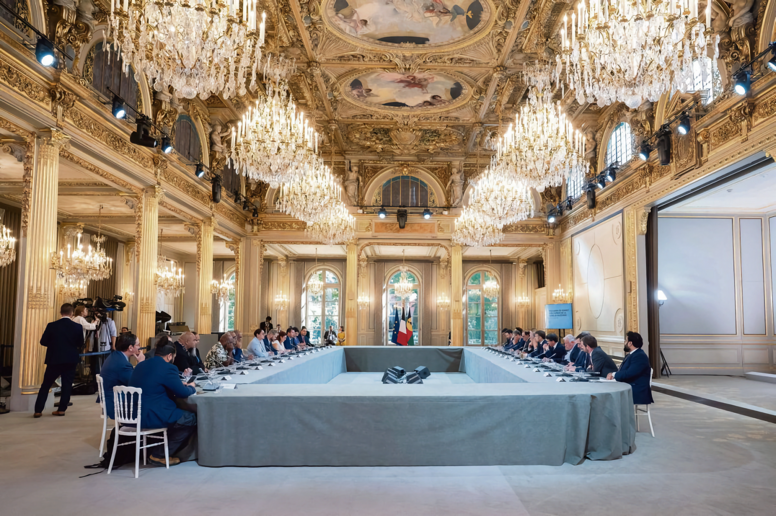 Une salle de réunion élégante avec des lustres en cristal, des draperies dorées et des colonnes ornementales, avec une longue table en forme de U entourée de personnes en réunion, et un drapeau français et un drapeau européen dans le fond.