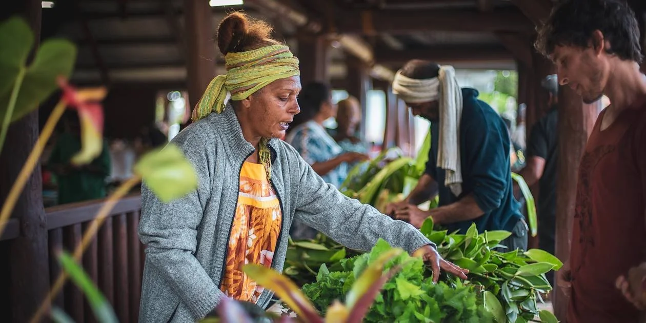 Une femme âgée vendant des plantes et des feuilles vertes à un marché ou une stand, entourée d'autres personnes également intéressées par les plantes.