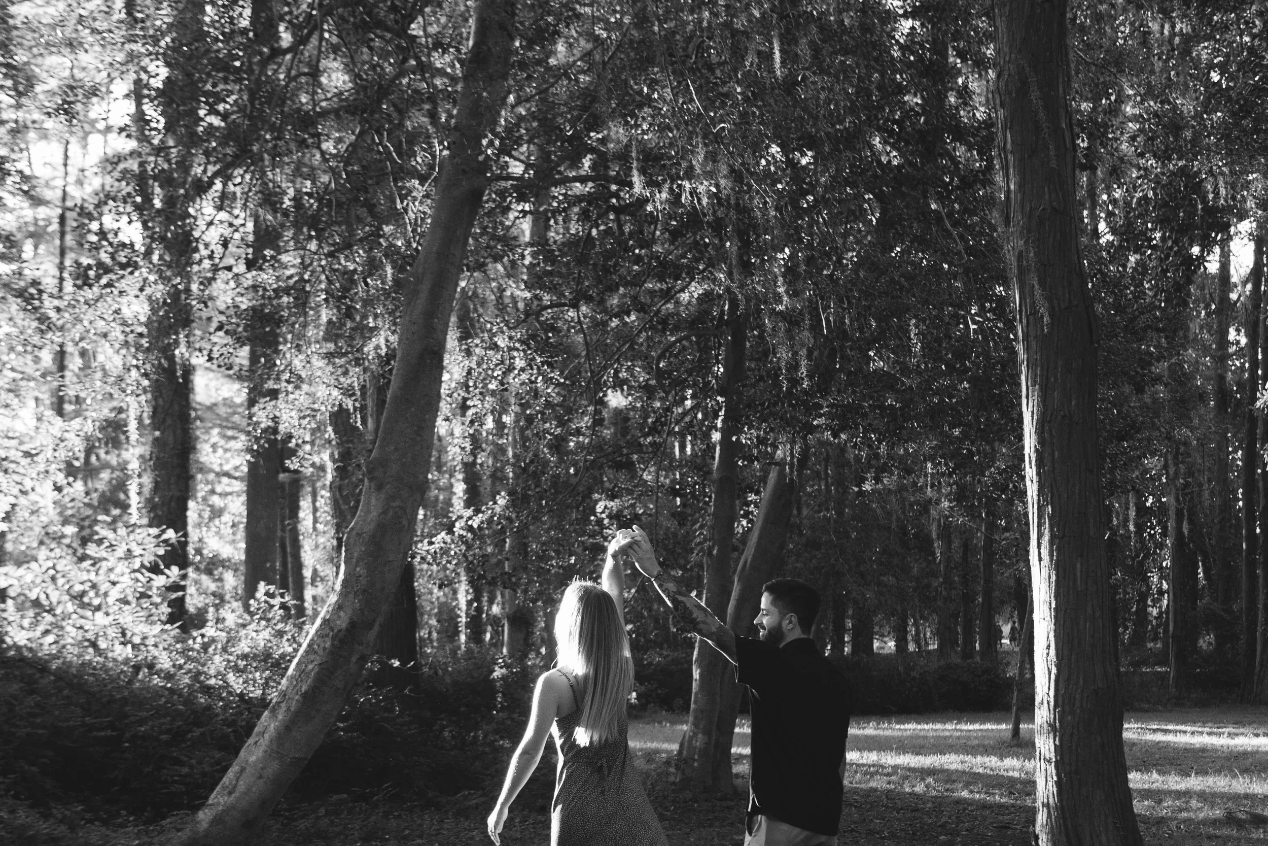 A black and white photo of a man and woman dancing in a forest, with the man twirling the woman by holding her hand up, surrounded by tall trees and dappled sunlight.
