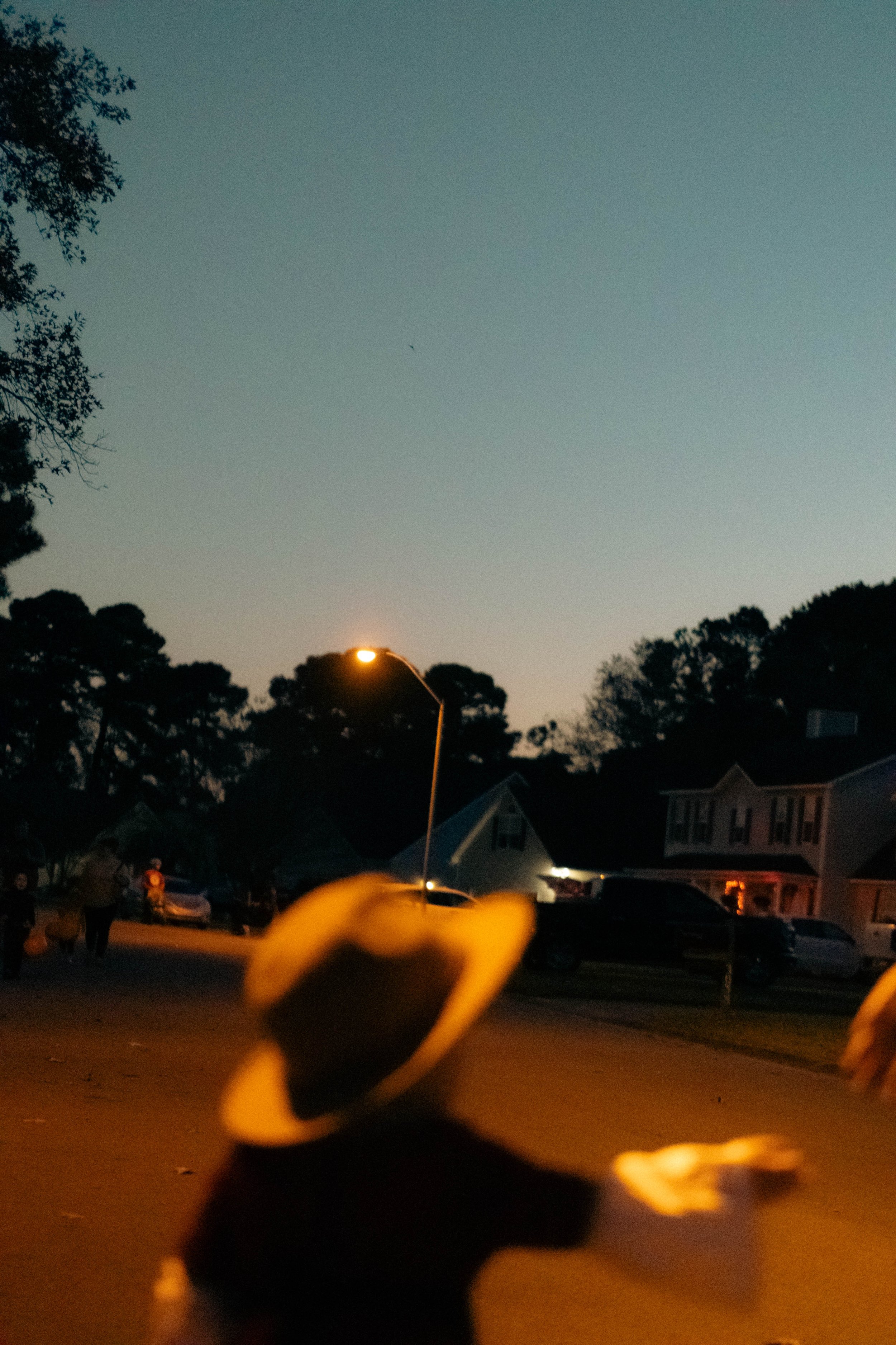 A photo taken at dusk or dawn showing a street with houses, parked cars, and trees. The sky is clear, and streetlights are on. There's a blurred figure or object in the foreground.