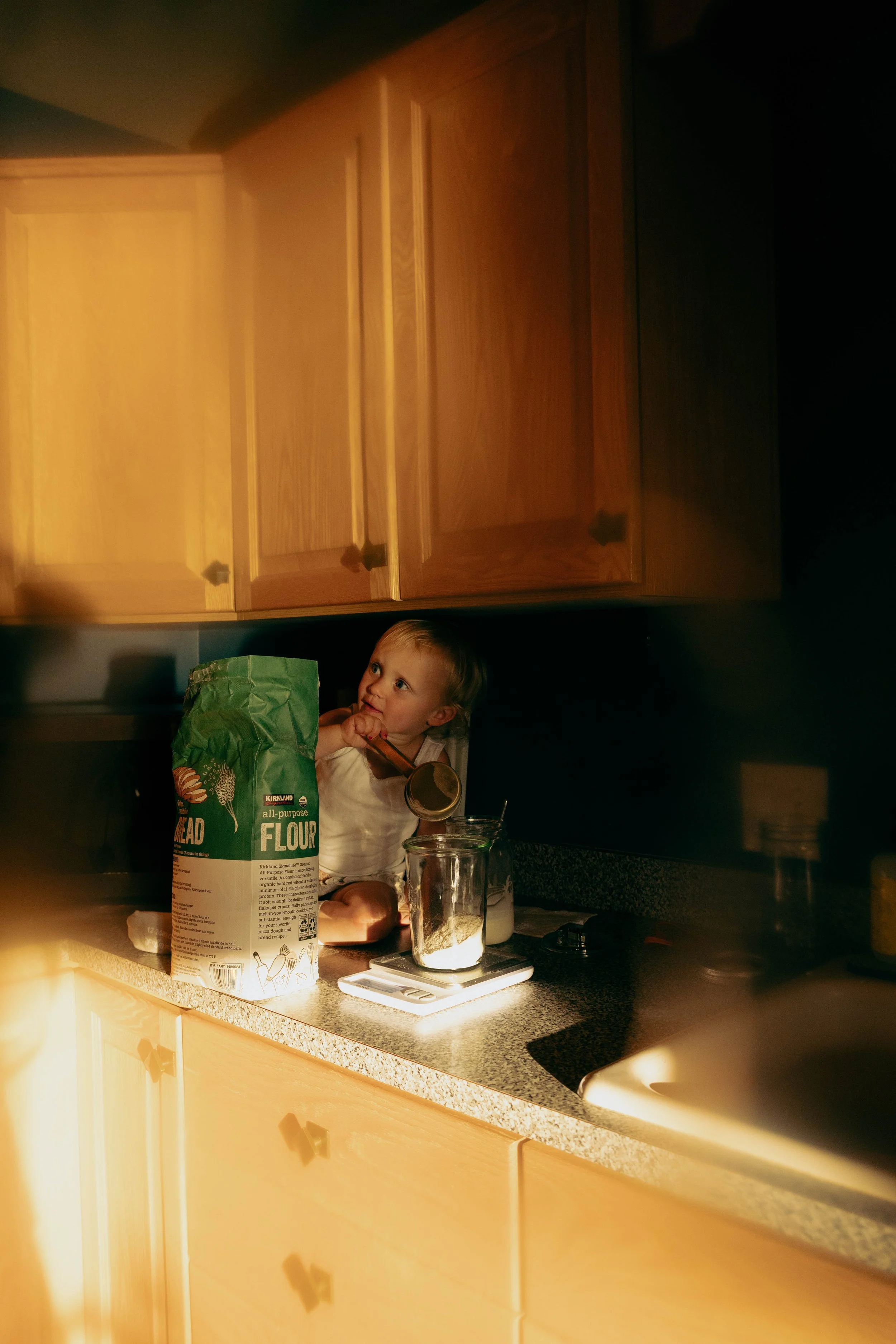 A young child standing on a kitchen counter in front of wooden cabinets, holding a wooden spoon and looking to the side, with a bag of all-purpose flour, measuring cups, and measuring scale around her.