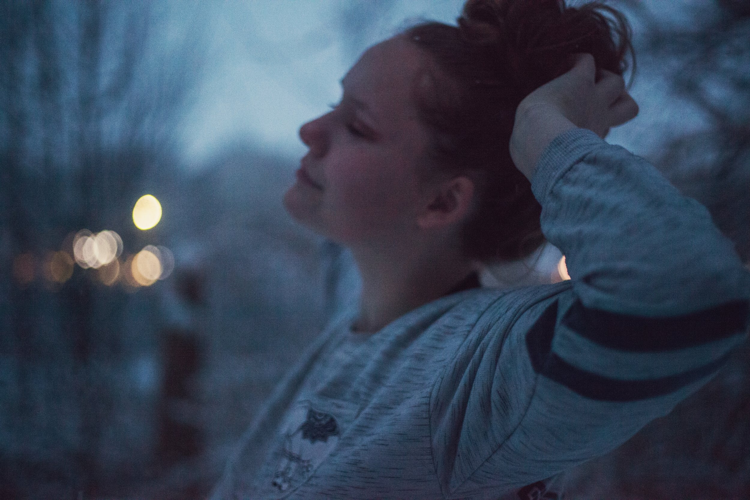 A young person with closed eyes, smiling while stretching their arms behind their head outdoors at dusk.