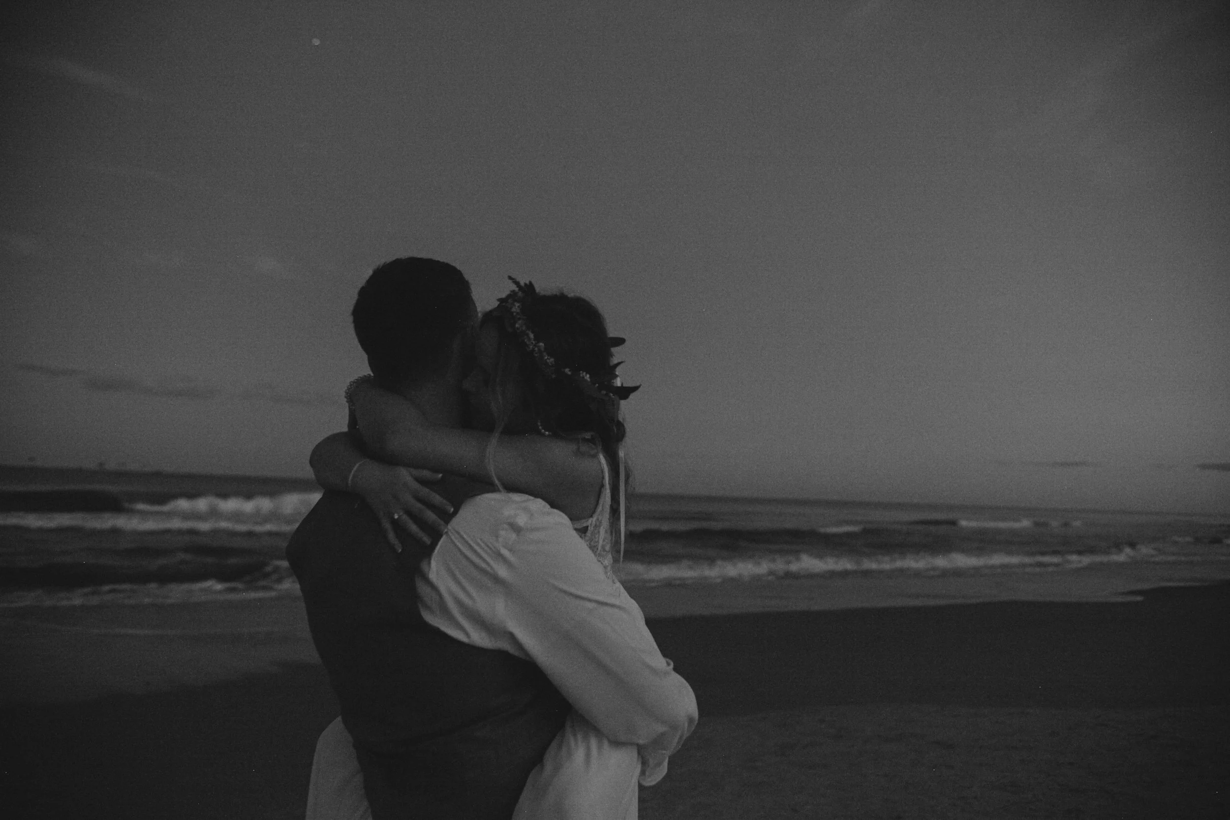 A freshly married couple embracing on the beach at dusk, with the ocean waves in the background, black and white photo.