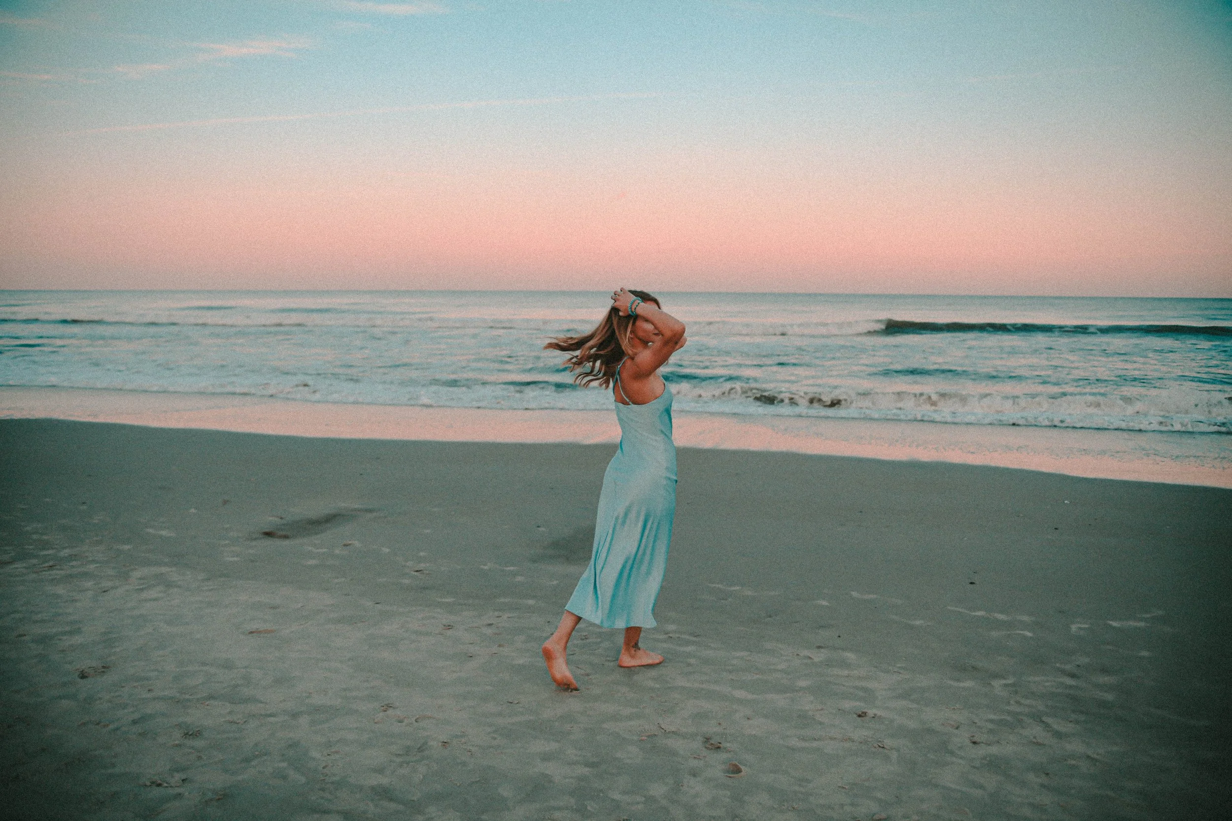 A woman in a blue dress walking barefoot on the beach during sunset, with the sea and pink sky in the background.