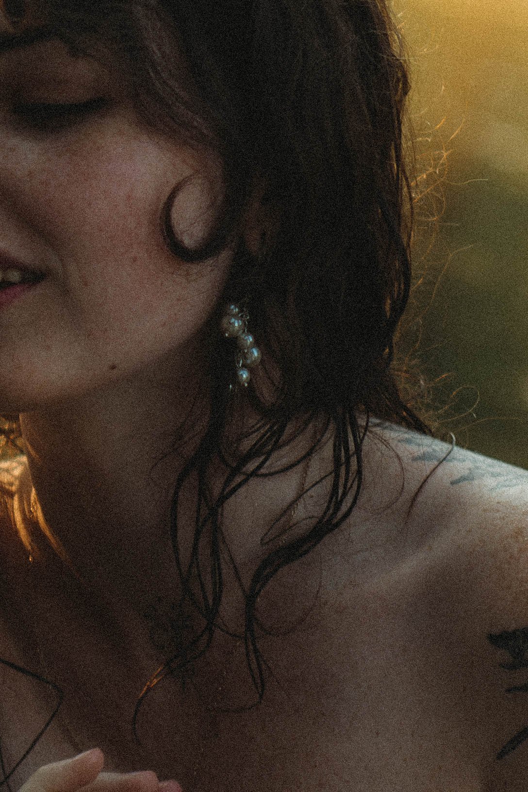 A close-up of a woman's face and shoulder, showing her curly dark hair, an earring with shimmering stones, and a tattoo on her shoulder. She has a slight smile.