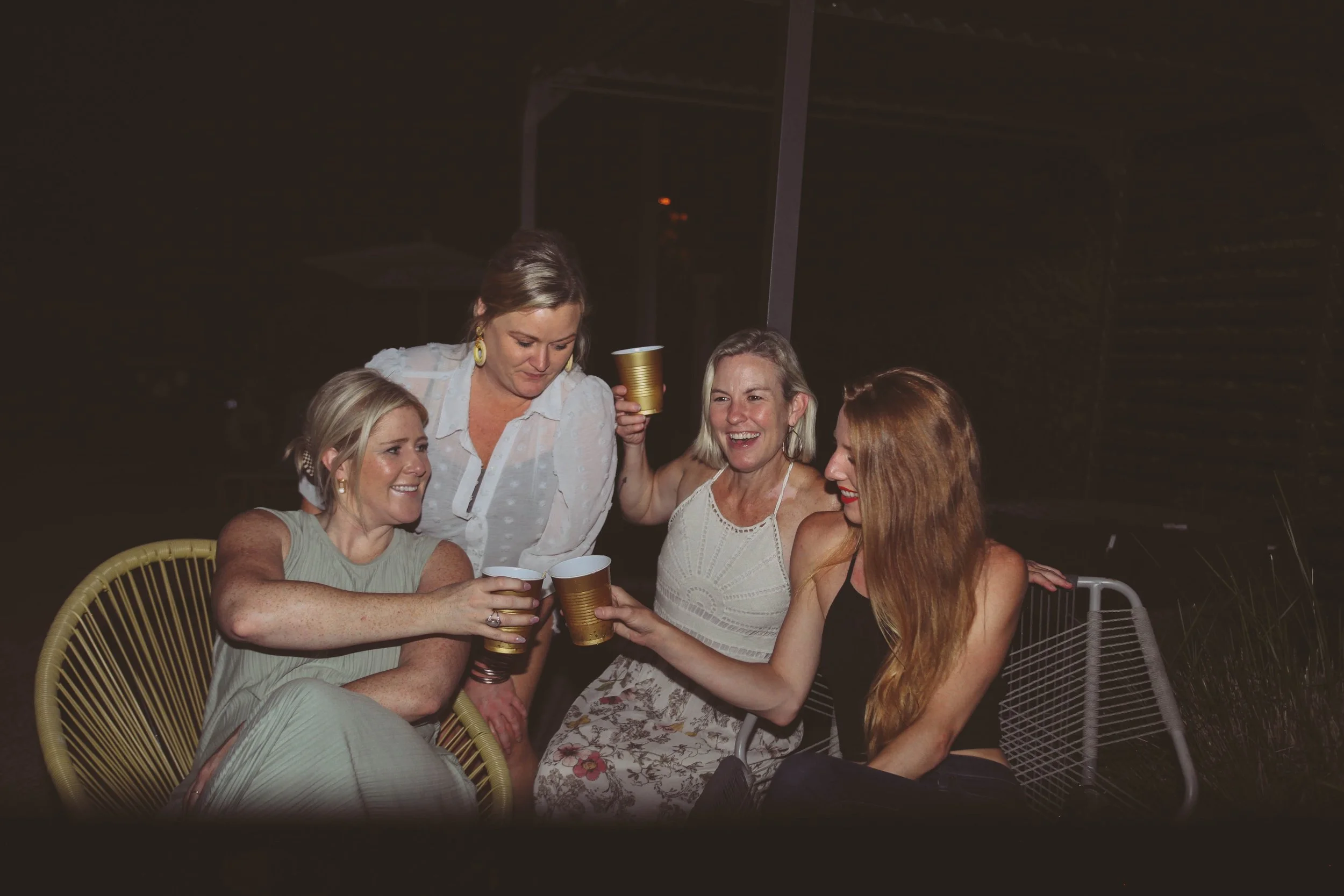 Four women enjoying drinks together at night, sitting on outdoor chairs and smiling.