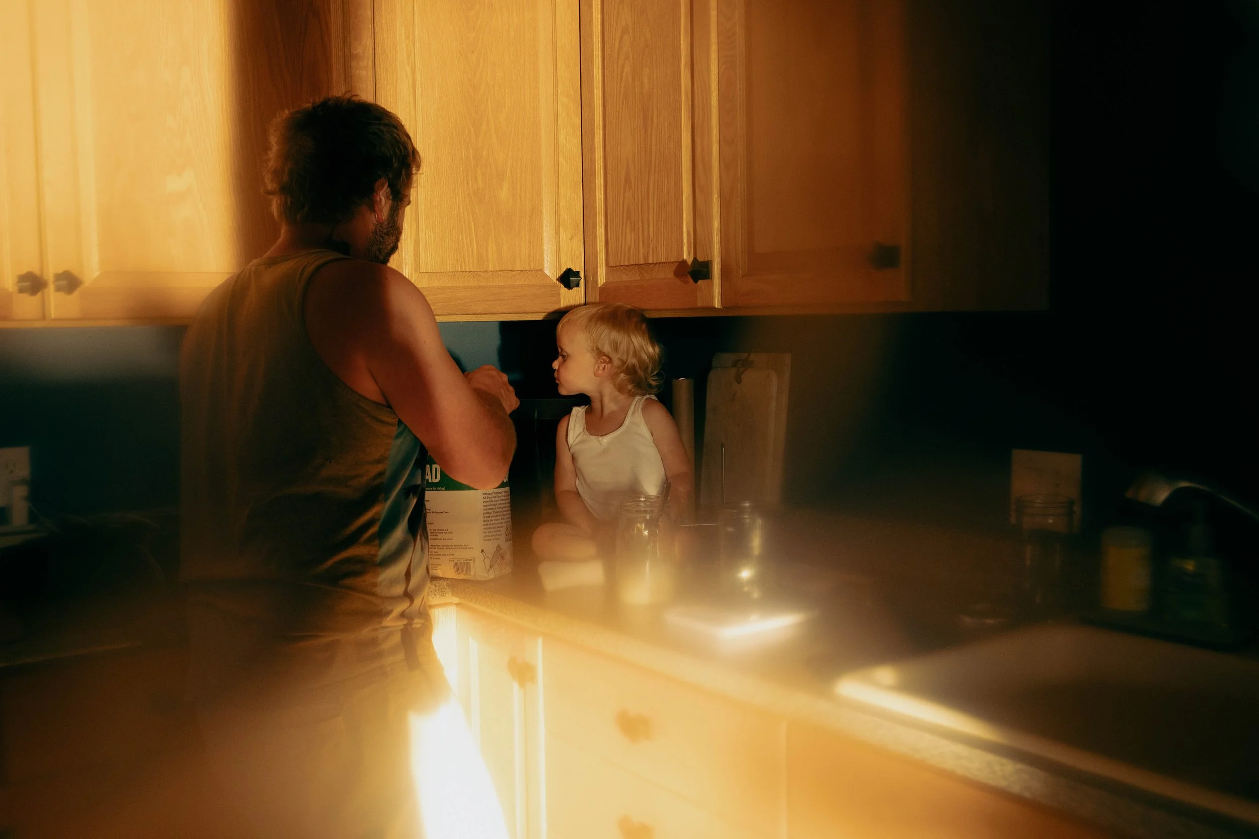 A man and a young girl sitting on a kitchen countertop, engaging in a conversation or activity, with warm lighting illuminating their faces and the wooden cabinets above.
