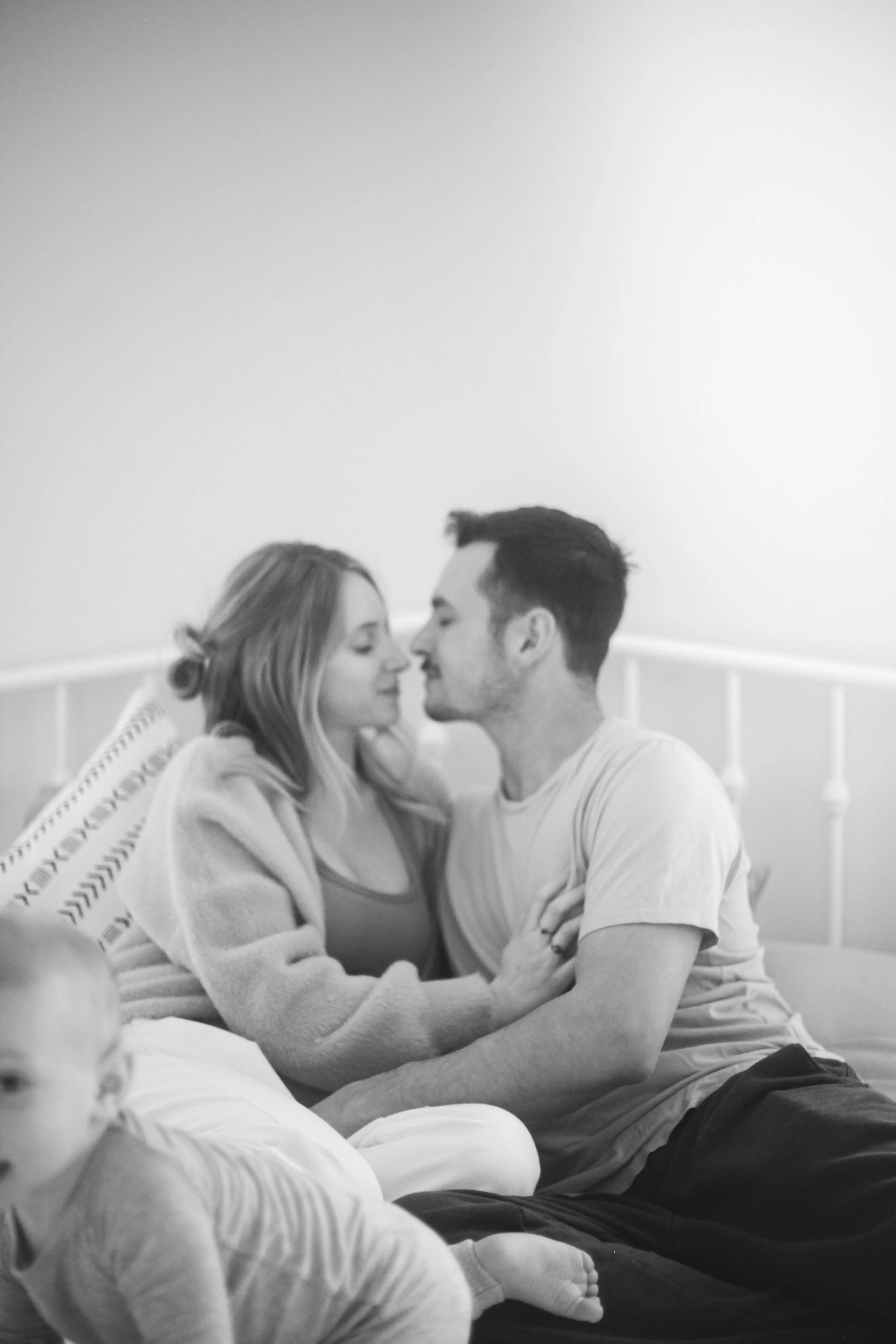 A black and white photo of a couple sitting on a bed, facing each other with noses touching, and a young child sitting in the foreground.